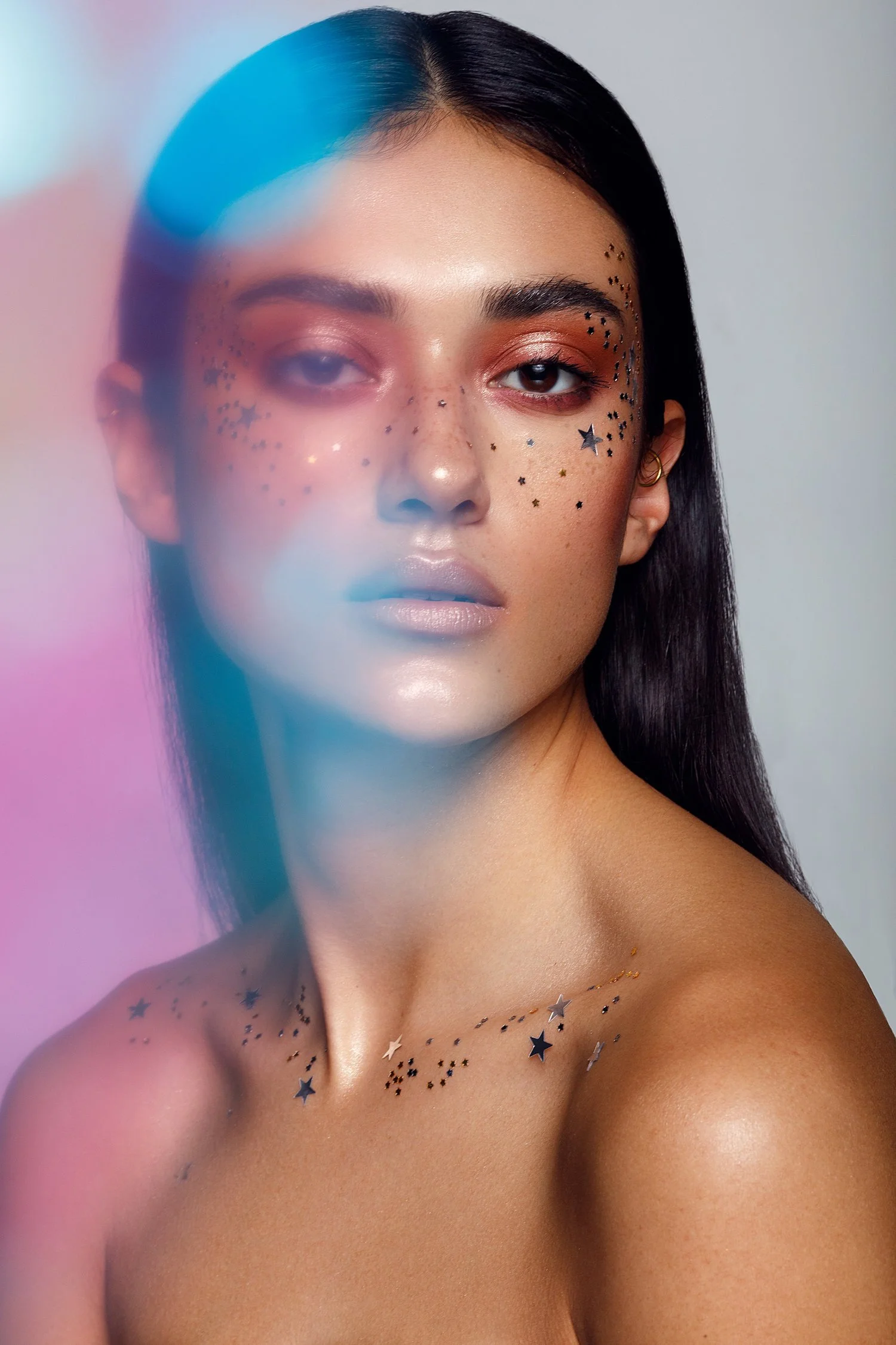 A woman with dark hair and makeup with star and dot face and shoulder glitter tattoos, looking at the camera with a neutral expression.