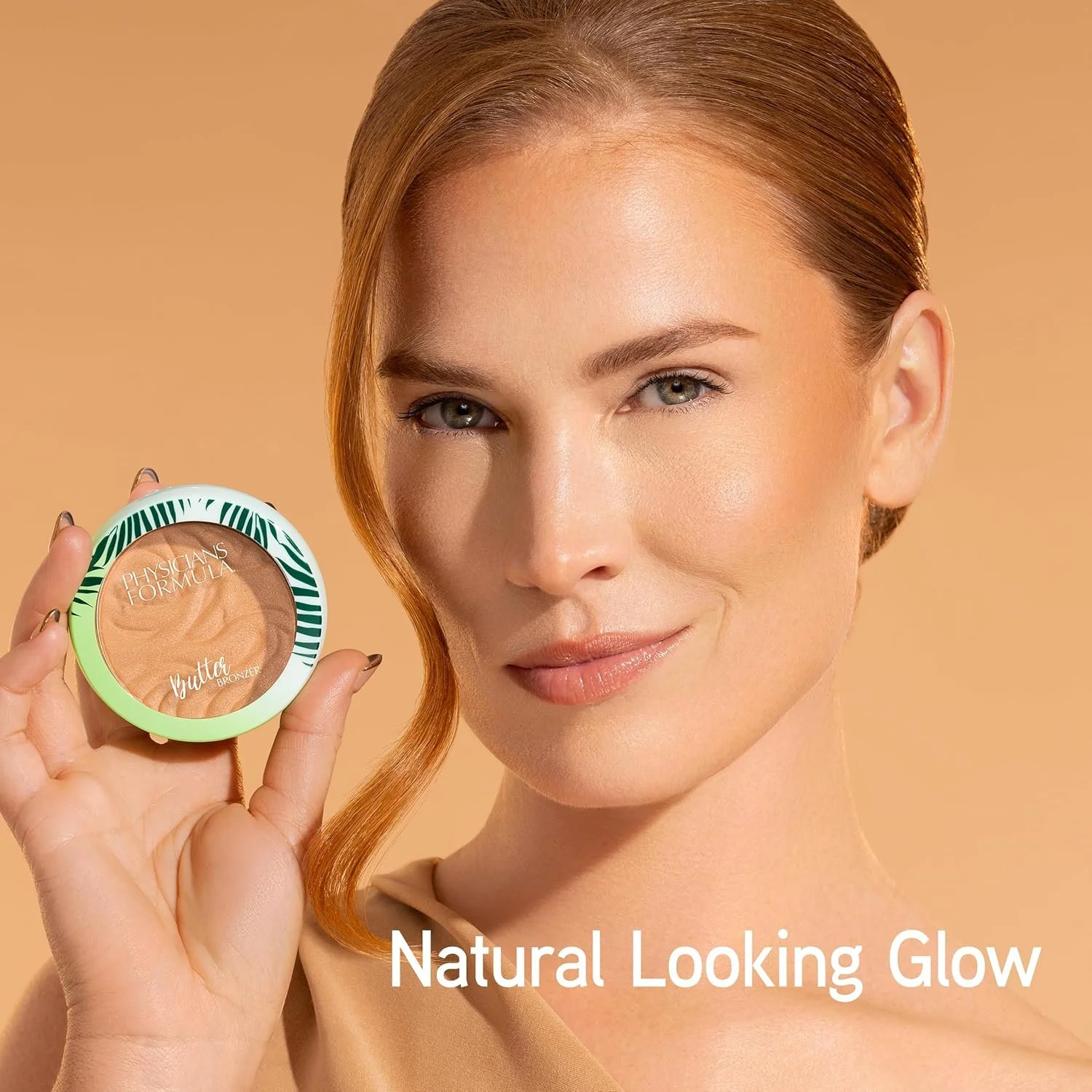 A woman with fair skin and light brown hair holds a bronzer compact labeled Physicans Formula Butter Bronze, smiling at the camera. The background is a neutral beige color, and the text "Natural Looking Glow" appears at the bottom of the image. Comme