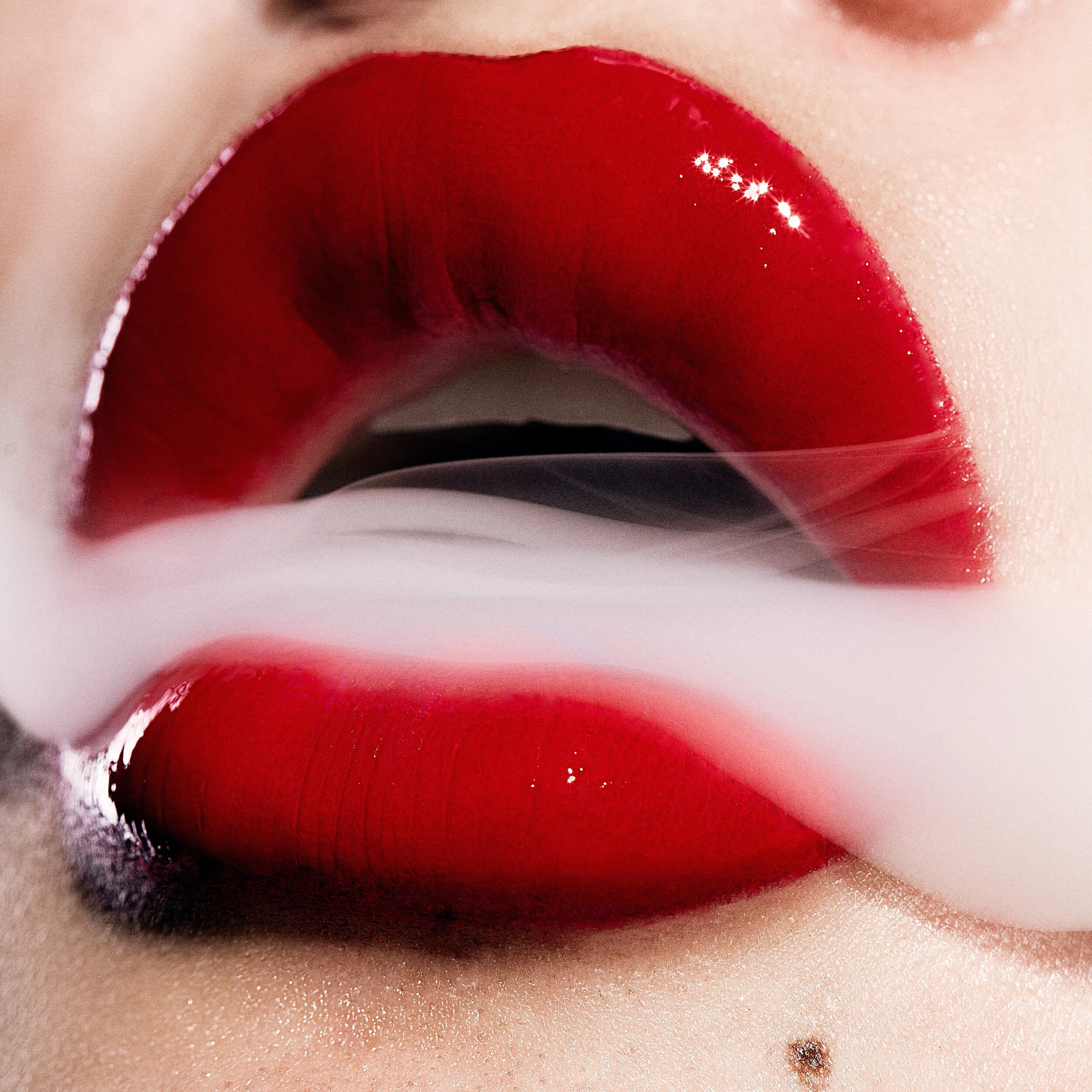 Close-up of lips painted in vibrant red lipstick, with a white mist or smoke partially covering the lips, and a small mole near the bottom right of the image. Beauty Photographer Los Angeles.