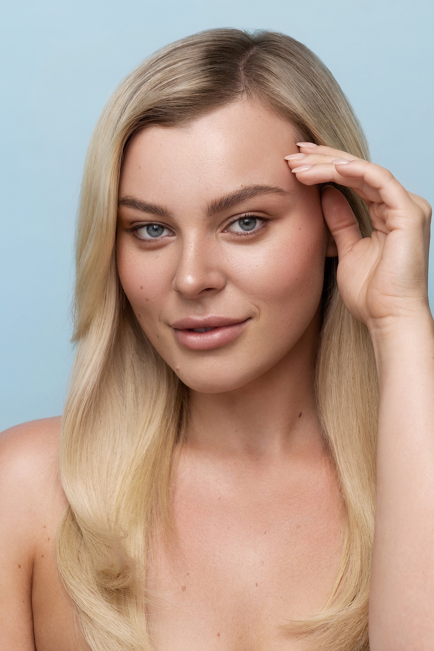 A young woman with long blonde hair and blue eyes, softly smiling, holding her hand to her forehead against a light blue background. Beauty Photographer Los Angeles.