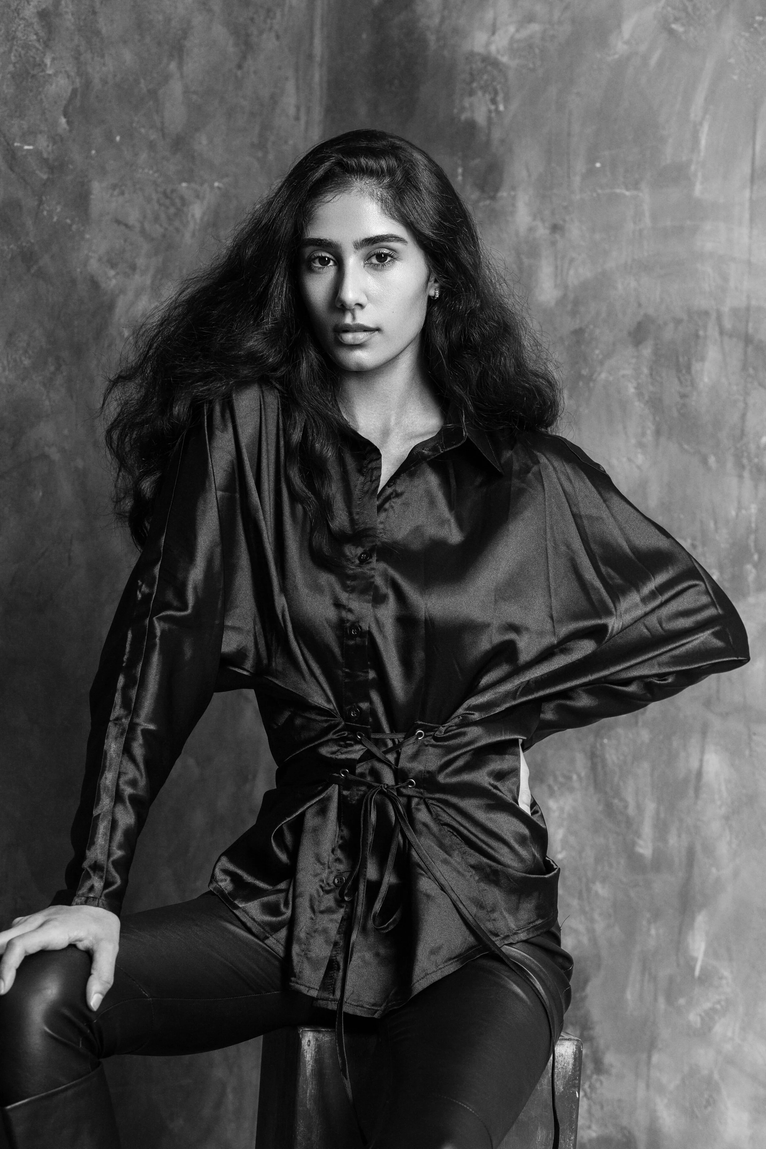 A young woman with long, curly hair poses sitting on a wooden stool in front of a textured gray background. She is wearing a shiny, dark satin button-up shirt and leather pants, looking directly at the camera.