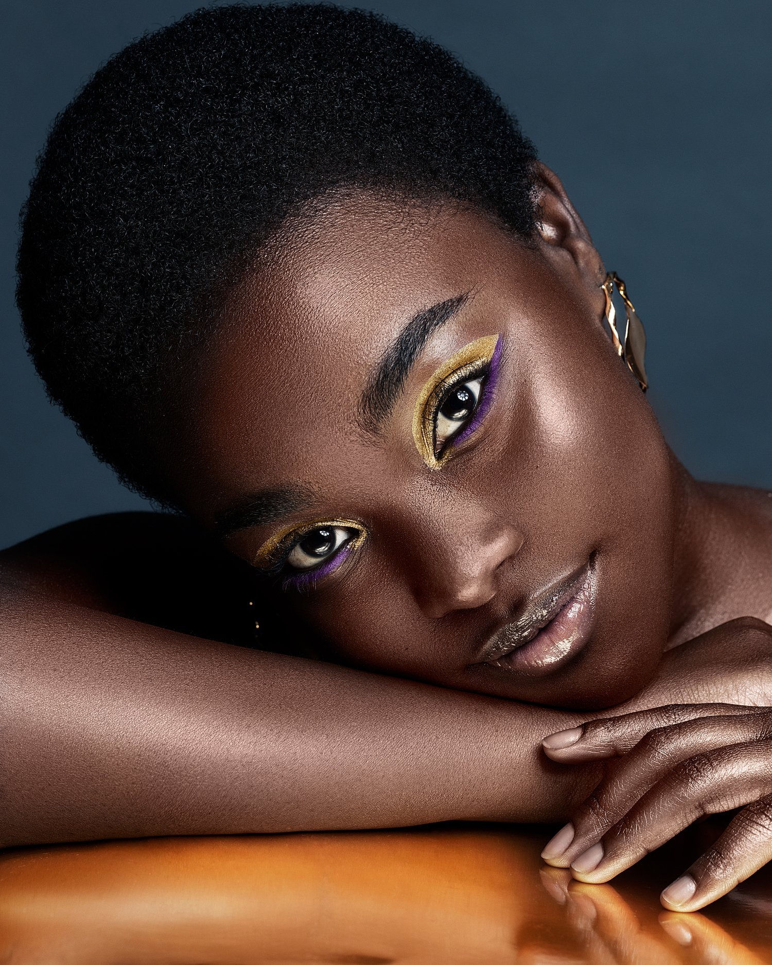 Close-up of a woman with short curly hair, wearing colorful eye makeup, gold and purple eyeshadow, and shiny metallic lipstick, resting her head on her folded arm. Beauty Photographer Los Angeles.