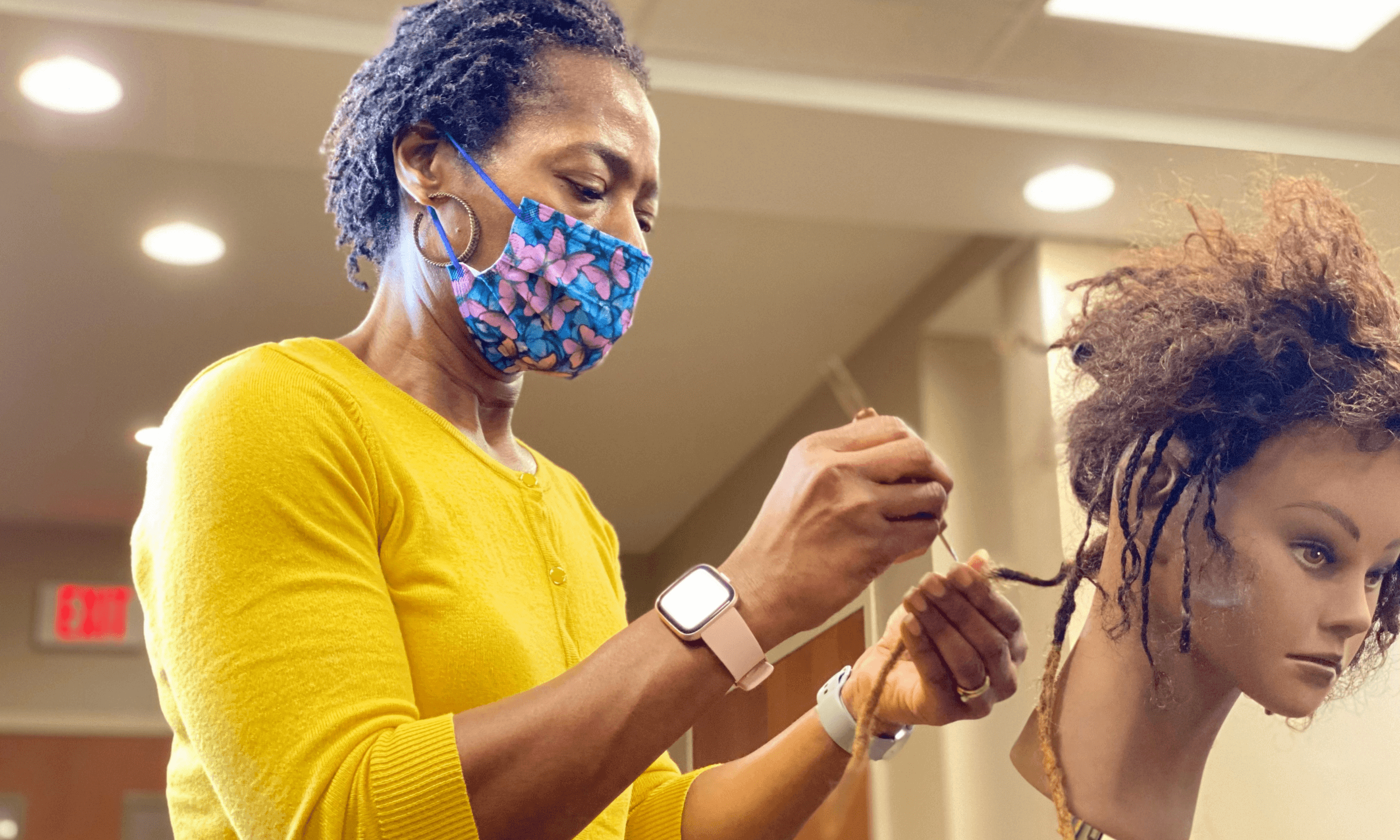 Woman with a colorful face mask practicing hairdressing on a mannequin with styled, textured hair.