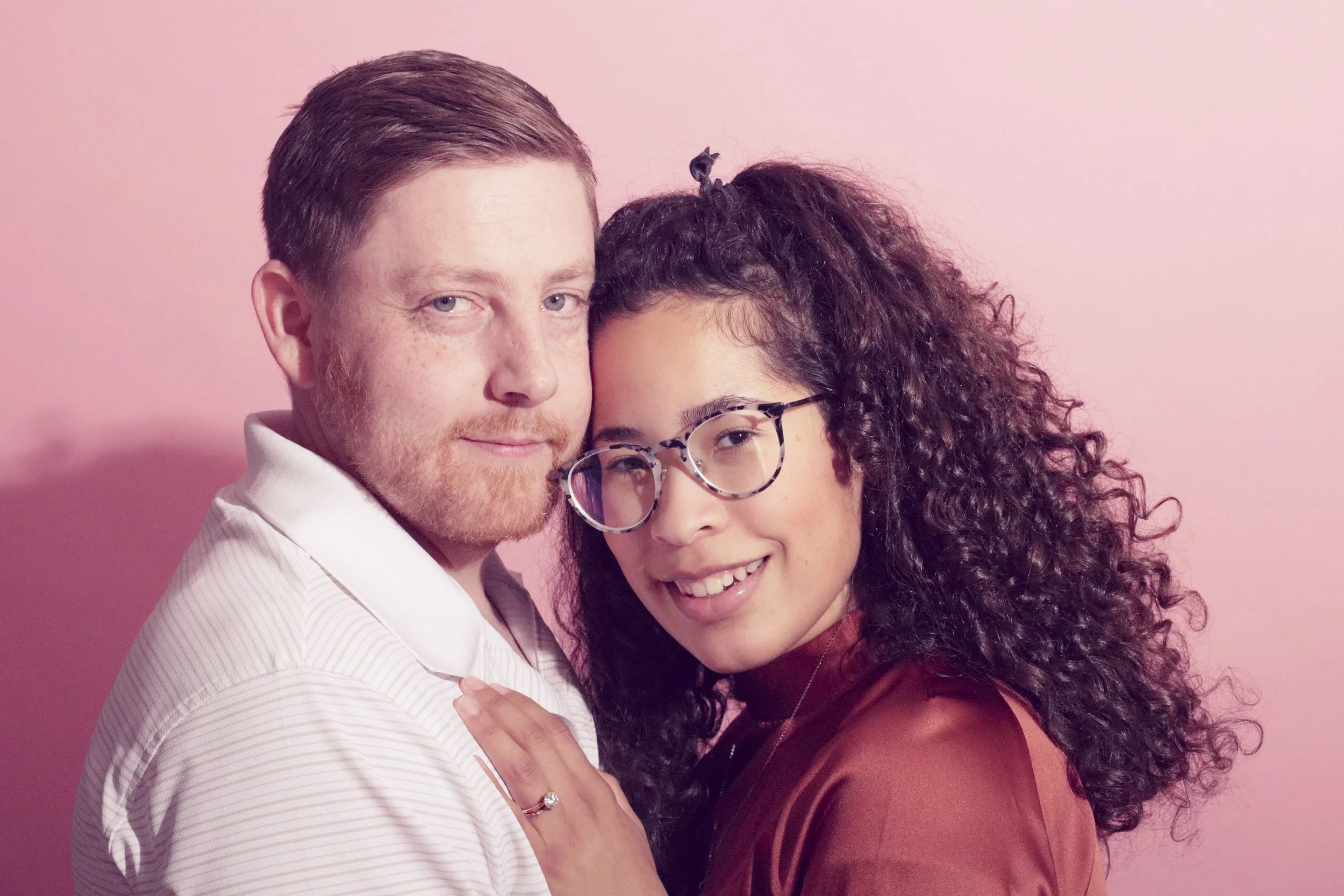 Portrait of a man and a woman hugging close facing the camera smiling with a pink background