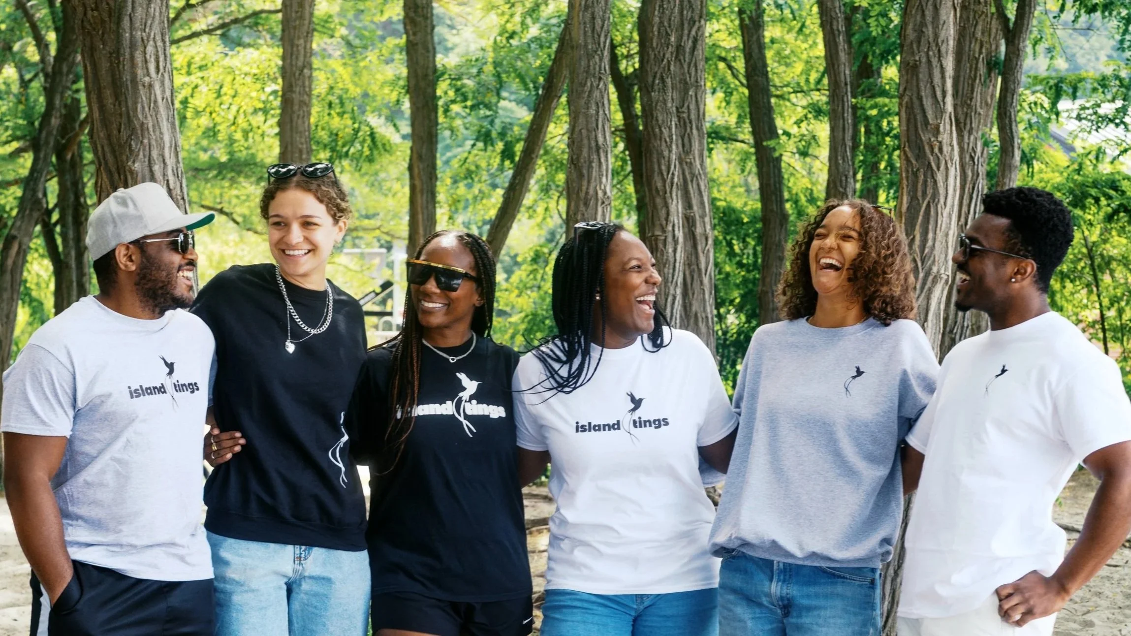Group of six diverse people smiling and laughing outdoors in a wooded area, wearing casual clothes with some sporting team shirts that read 'island kings' and a bird logo.