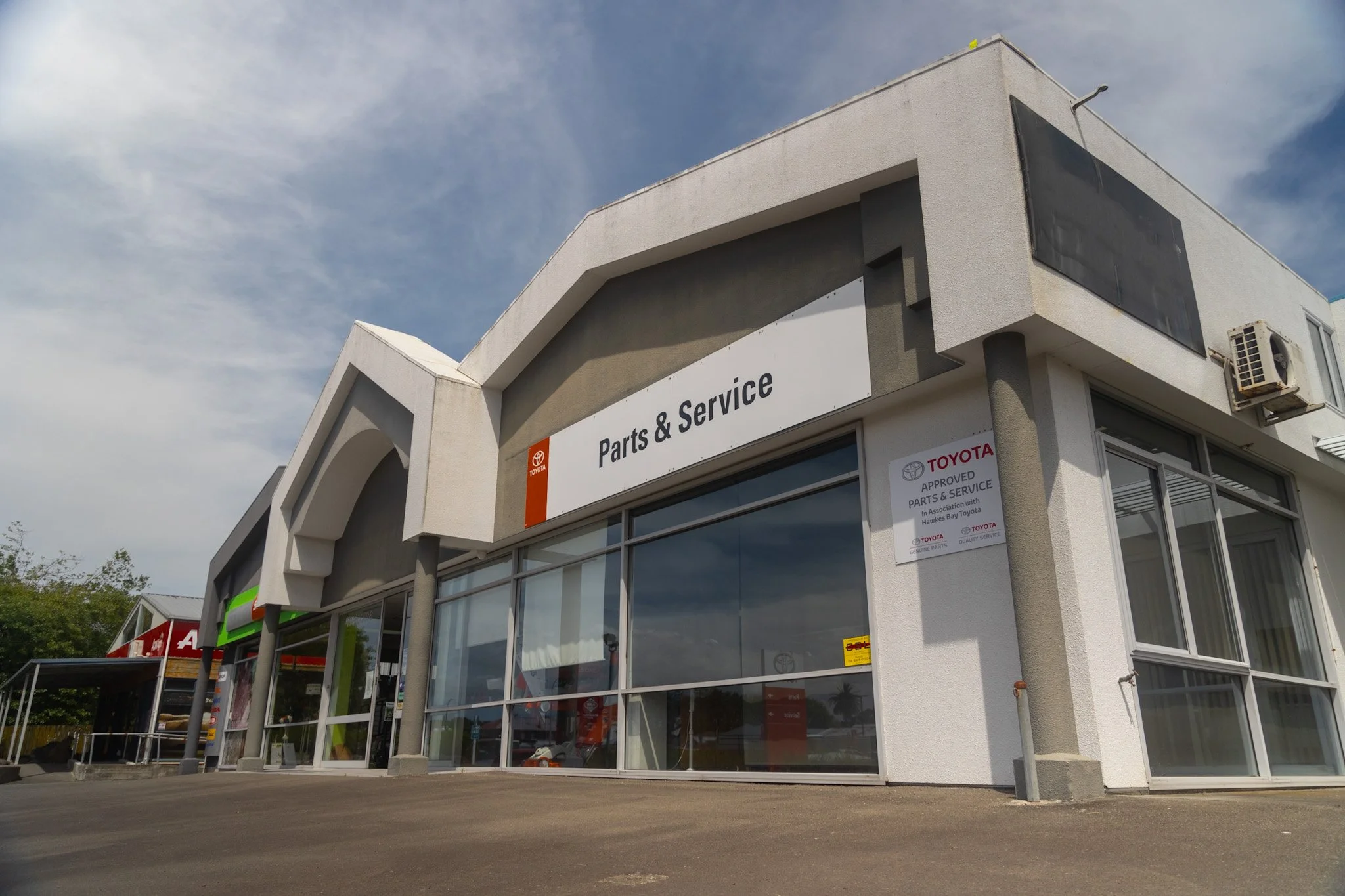 Exterior view of a building housing a parts and service store with large windows and a sign reading 'Parts & Service'. The sky is partly cloudy, and the parking lot is empty.