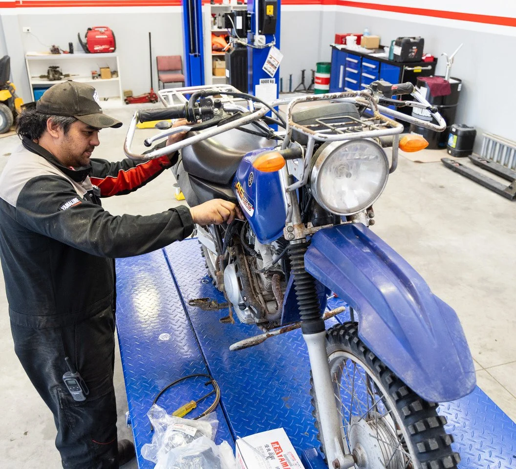 A mechanic working on a blue off-road motorcycle inside a garage or workshop with tools and equipment in the background.