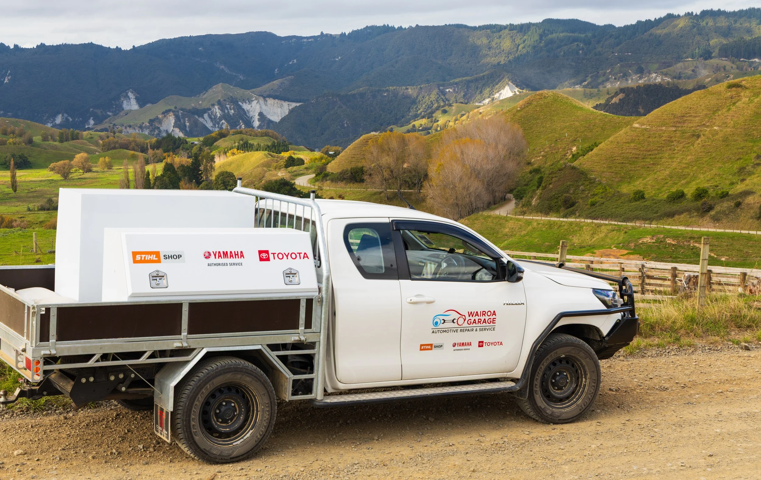 White pickup truck with company logos parked on a dirt road in front of green rolling hills and mountains under a cloudy sky.