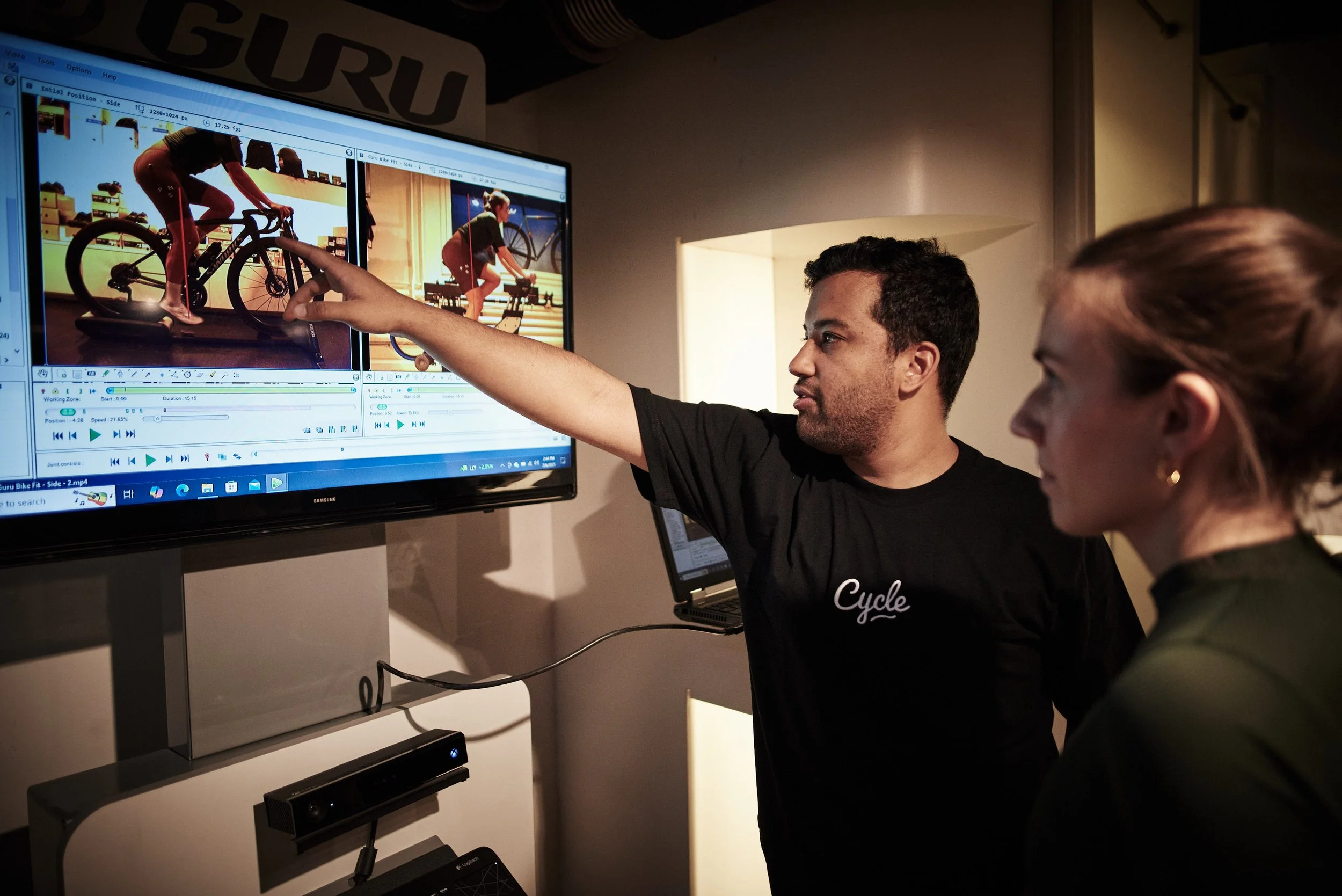 A man is pointing at a monitor displaying videos of a person cycling on a stationary bike, with a woman observing.
