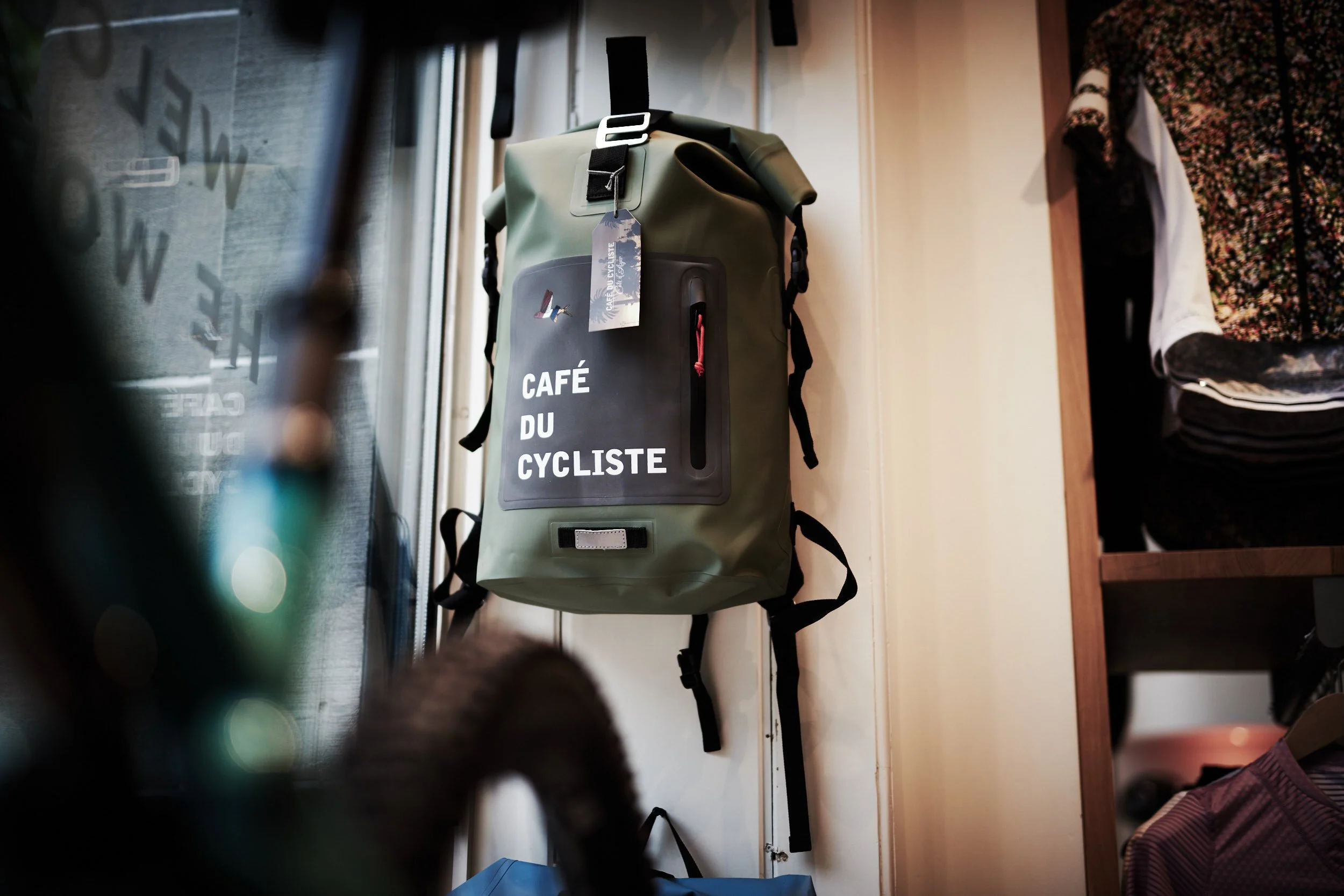 A green backpack with the words 'Café du Cycliste' hangs on a wall, with clothes on a shelf next to it.