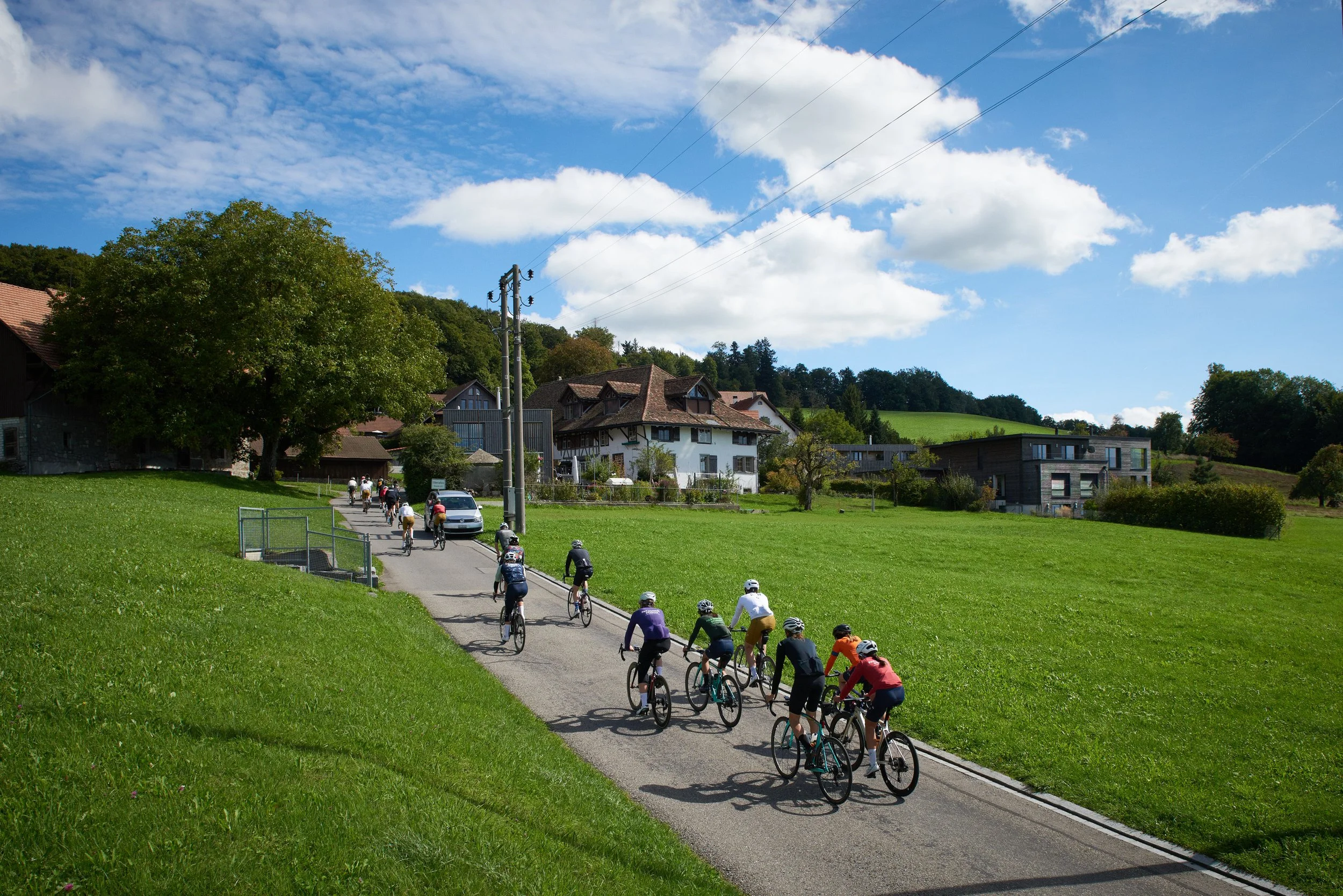 Group of cyclists riding on a paved road in a rural area with greenery and houses under a blue sky with clouds.