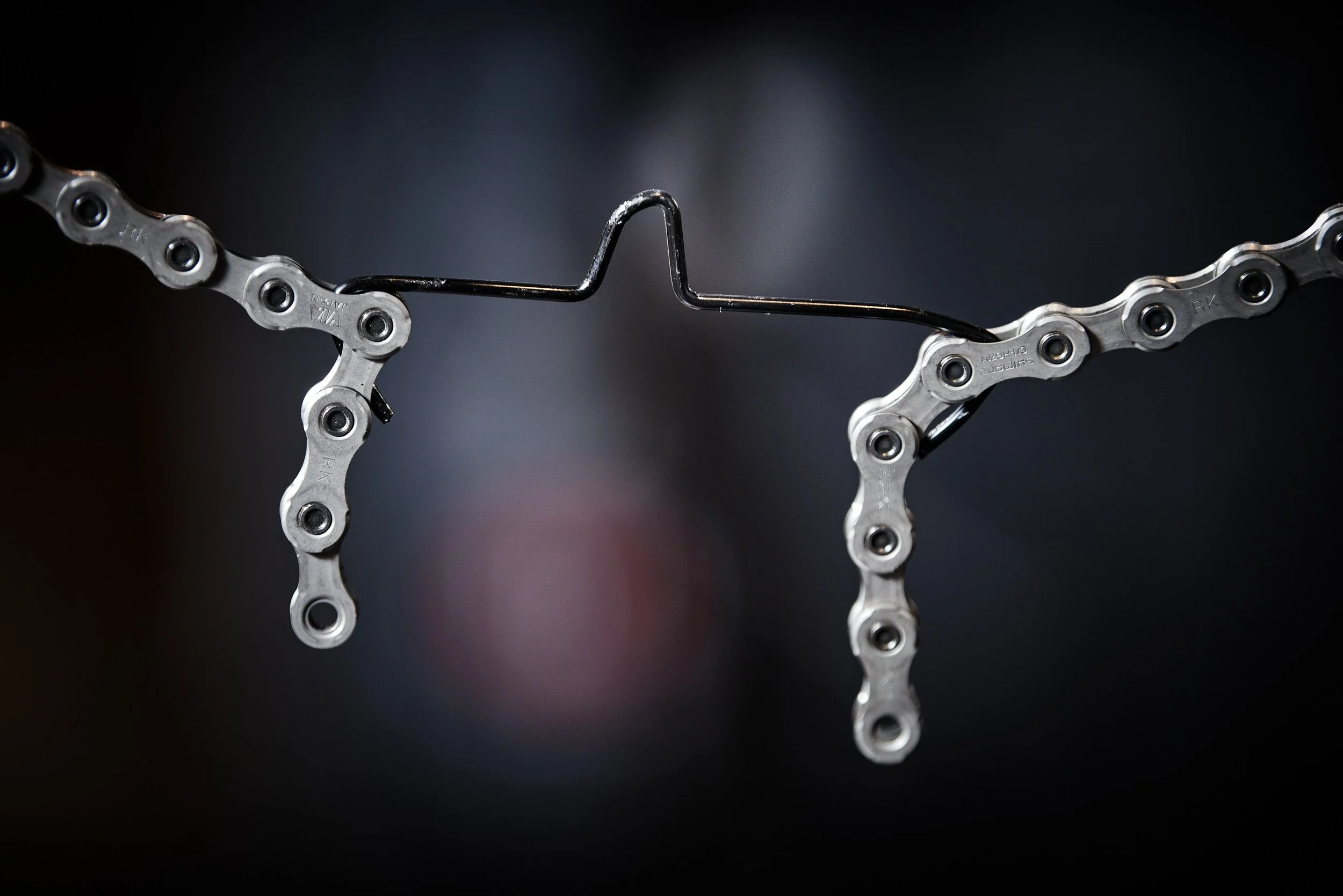Close-up of a bicycle chain with a black metal wire hanging through the middle, against a dark blurred background.