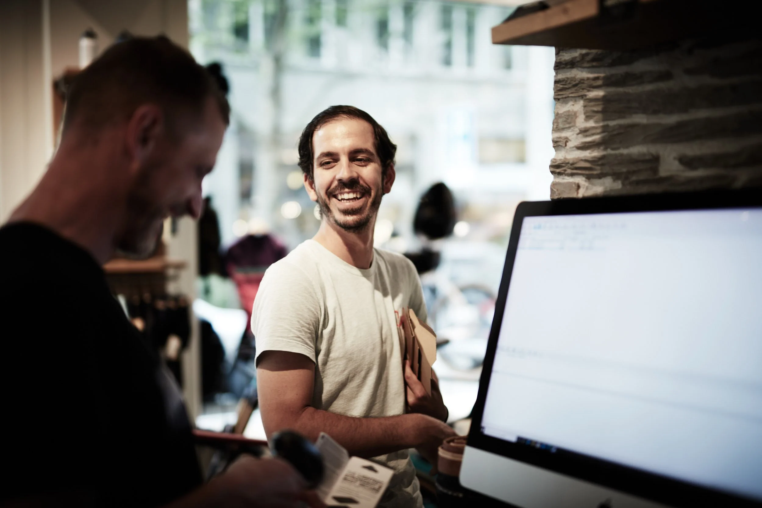 A man with a beard and short dark hair smiling and holding a package inside a store, while another person appears to be using a scanner or phone. The scene has natural light coming through a window.