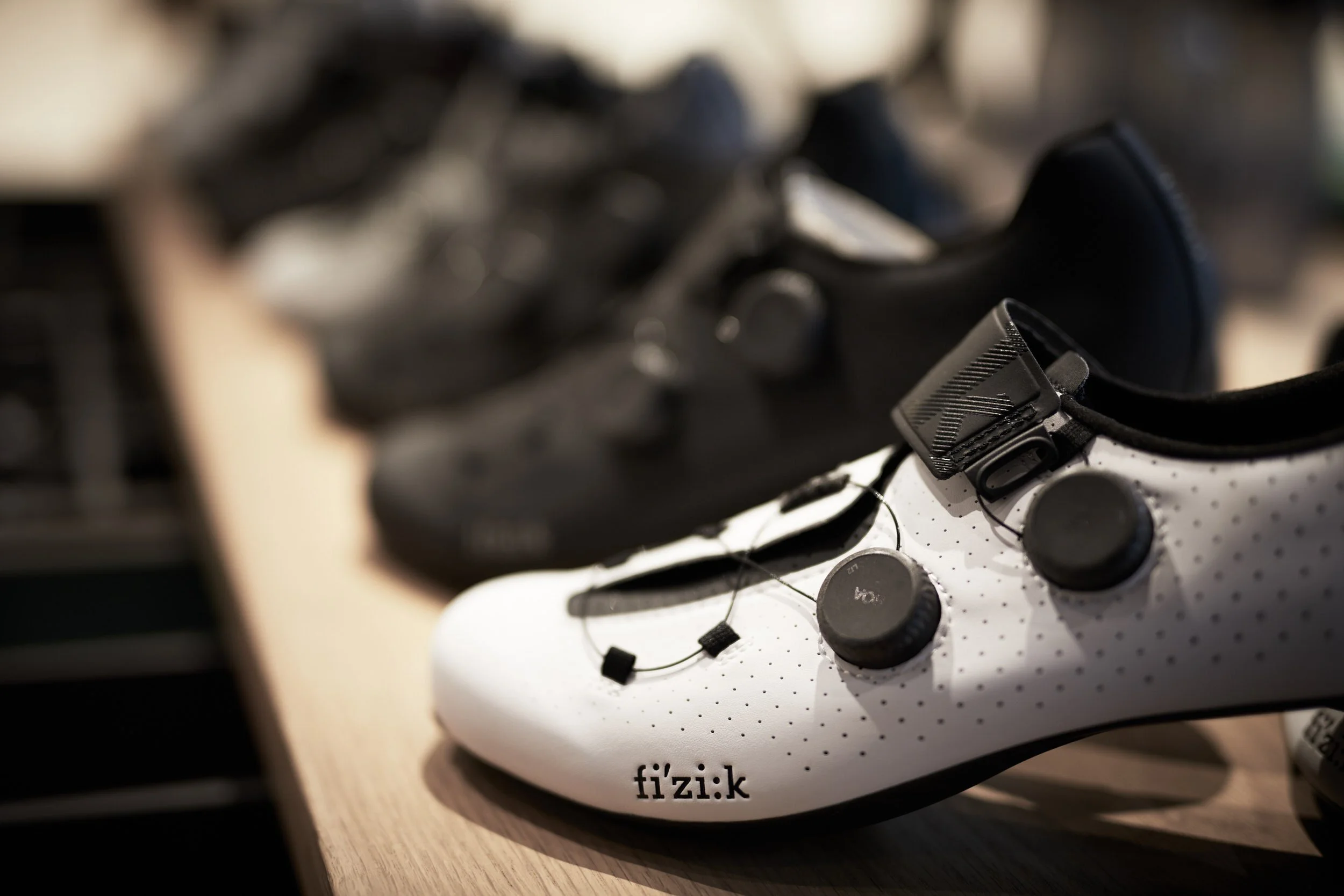 A white cycling shoe with black accents and the word 'fizik' on the toe, placed on a wooden surface with a pair of black cycling shoes in the background.