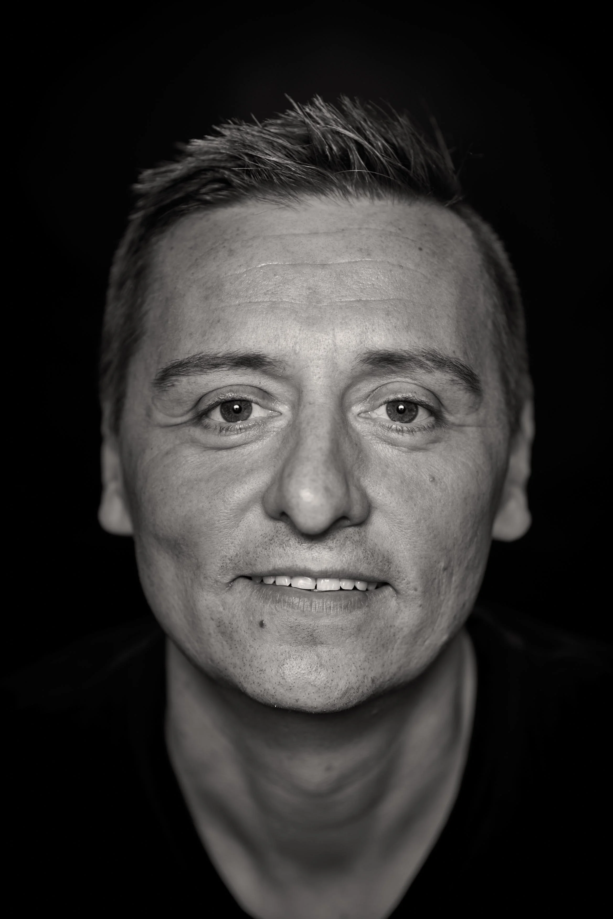 Black and white portrait of a middle-aged man with short hair, smiling slightly, against a dark background.