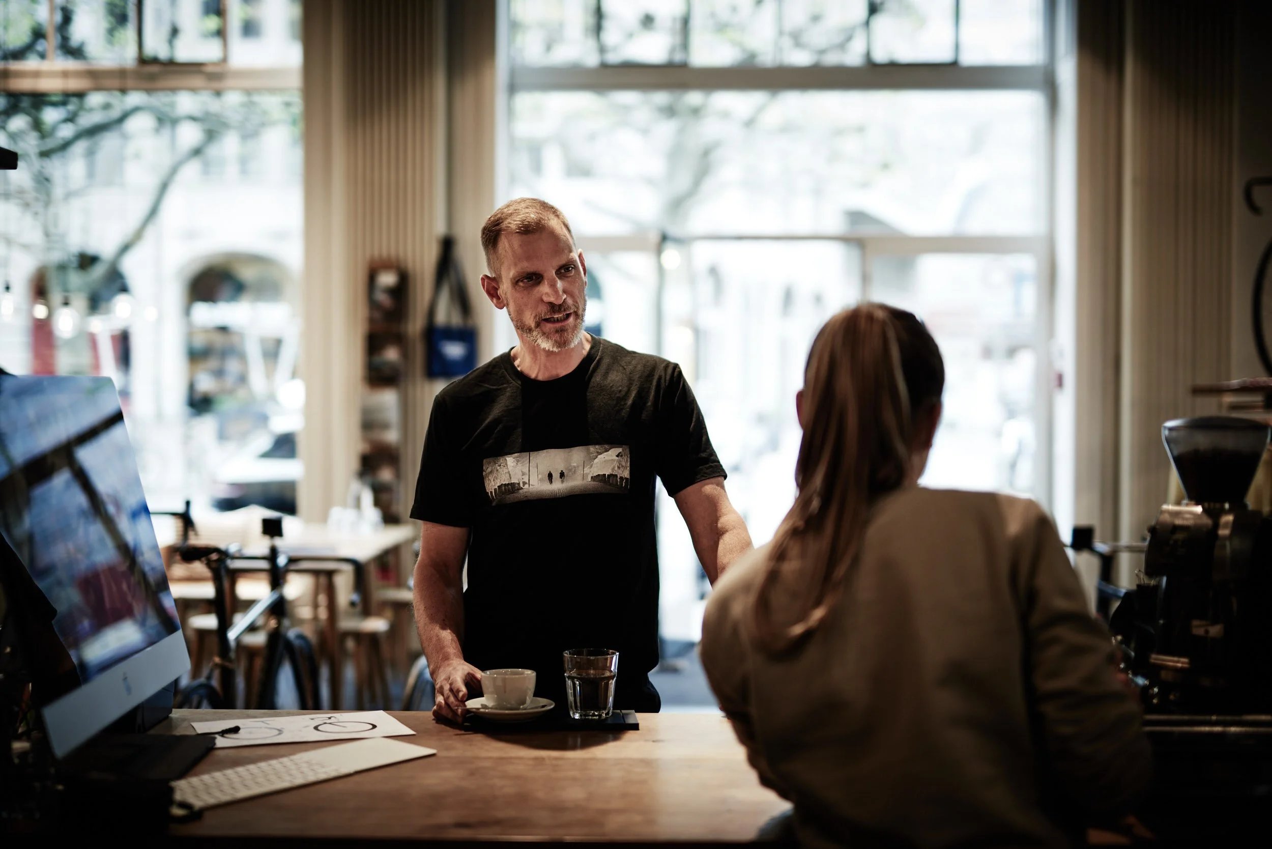 A barista with a beard and black t-shirt talking to a customer at a coffee shop counter, holding a coffee cup and spoon, with a computer and coffee machine on the counter.