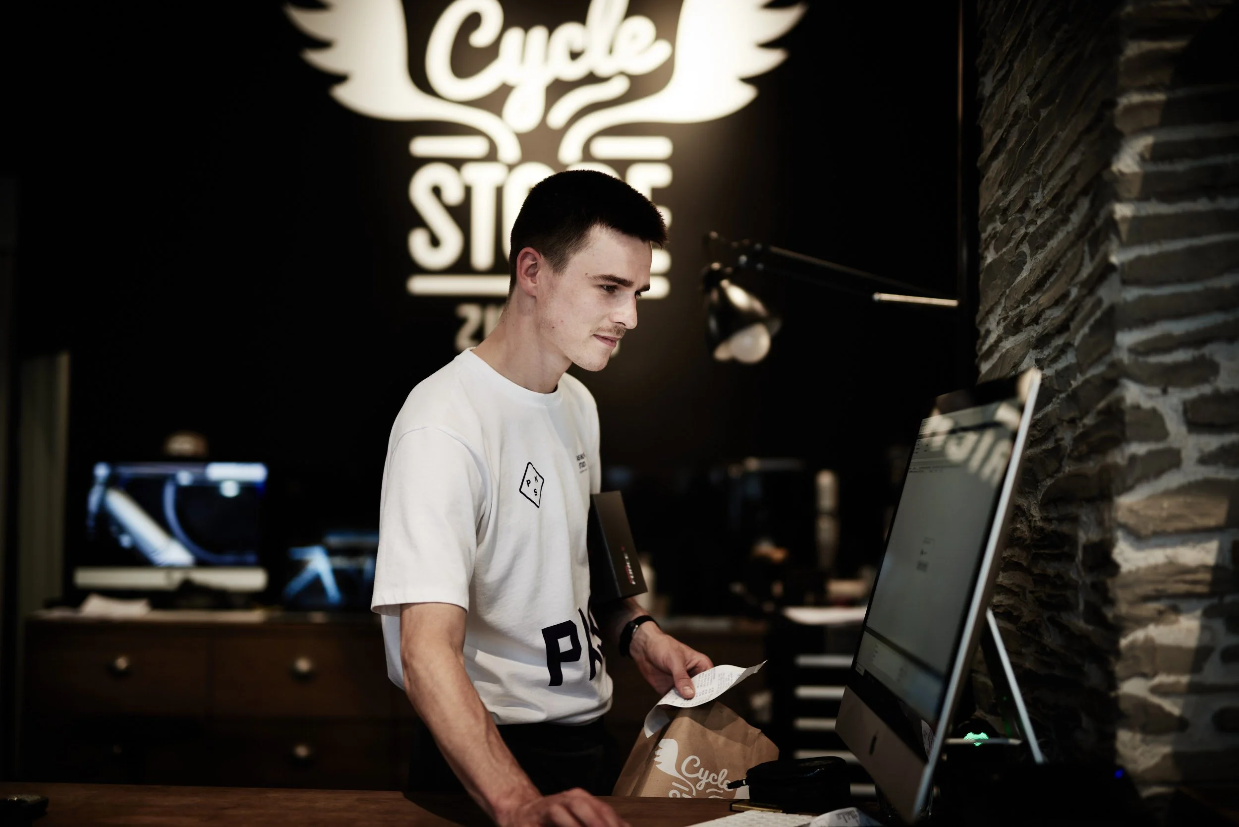 A young man at a counter in a cafe or restaurant, holding a receipt and a paper bag, looking at a computer monitor. The background features a black wall with a illuminated sign reading 'Cycle Store'.