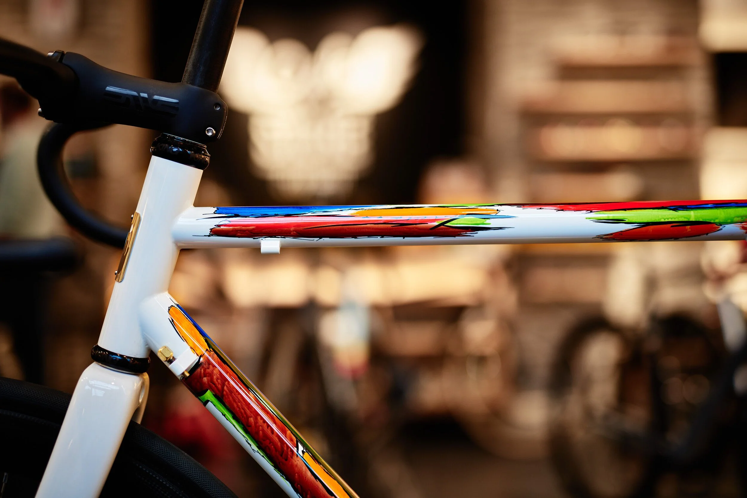 Close-up of a colorful bicycle frame with a white base color and abstract multicolor paint streaks, focused on the top tube and part of the seat tube.