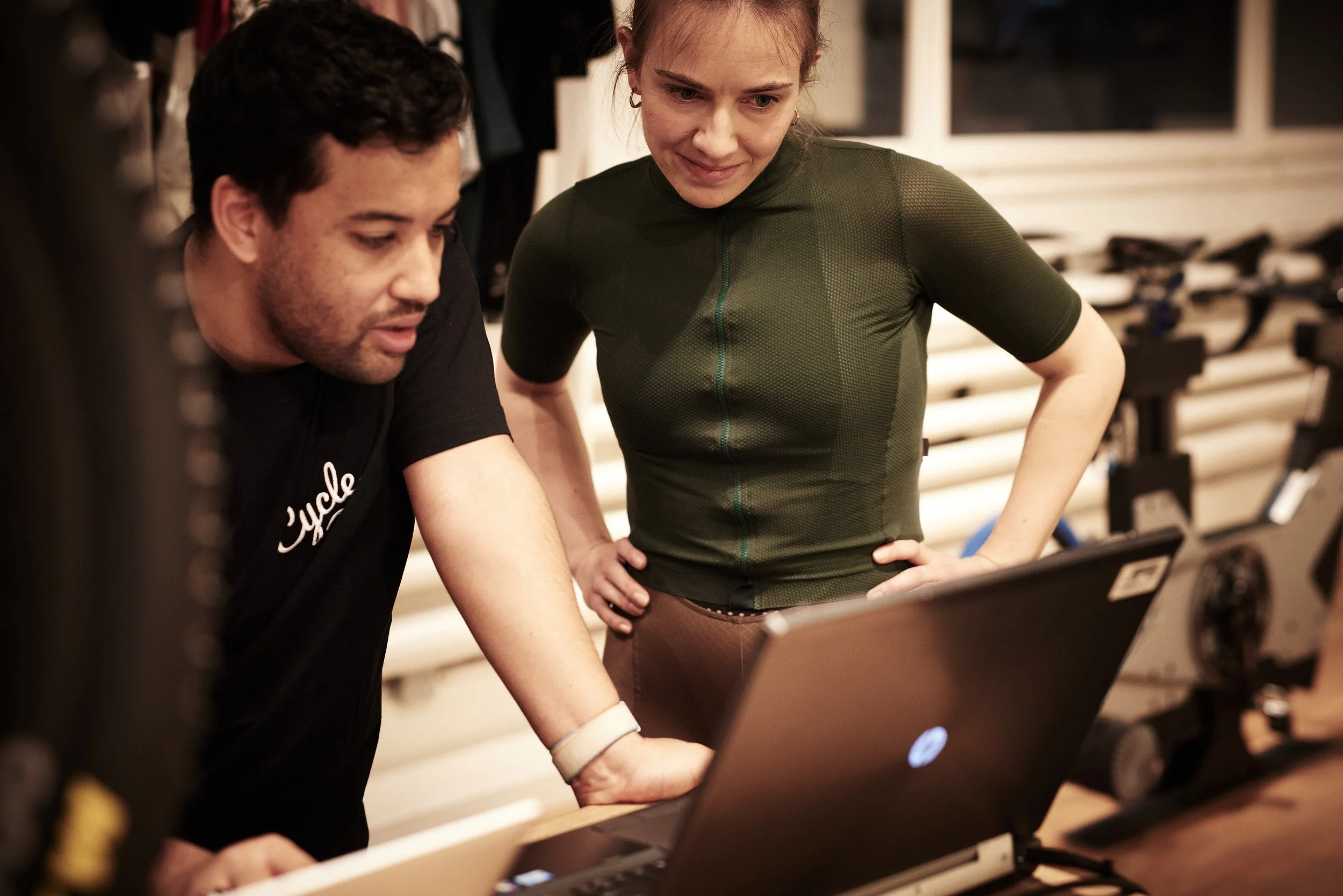 Two people, a man and a woman, are standing closely together looking at a laptop screen. They appear to be discussing or reviewing something, possibly in a gym or workout setting, with exercise equipment visible in the background.
