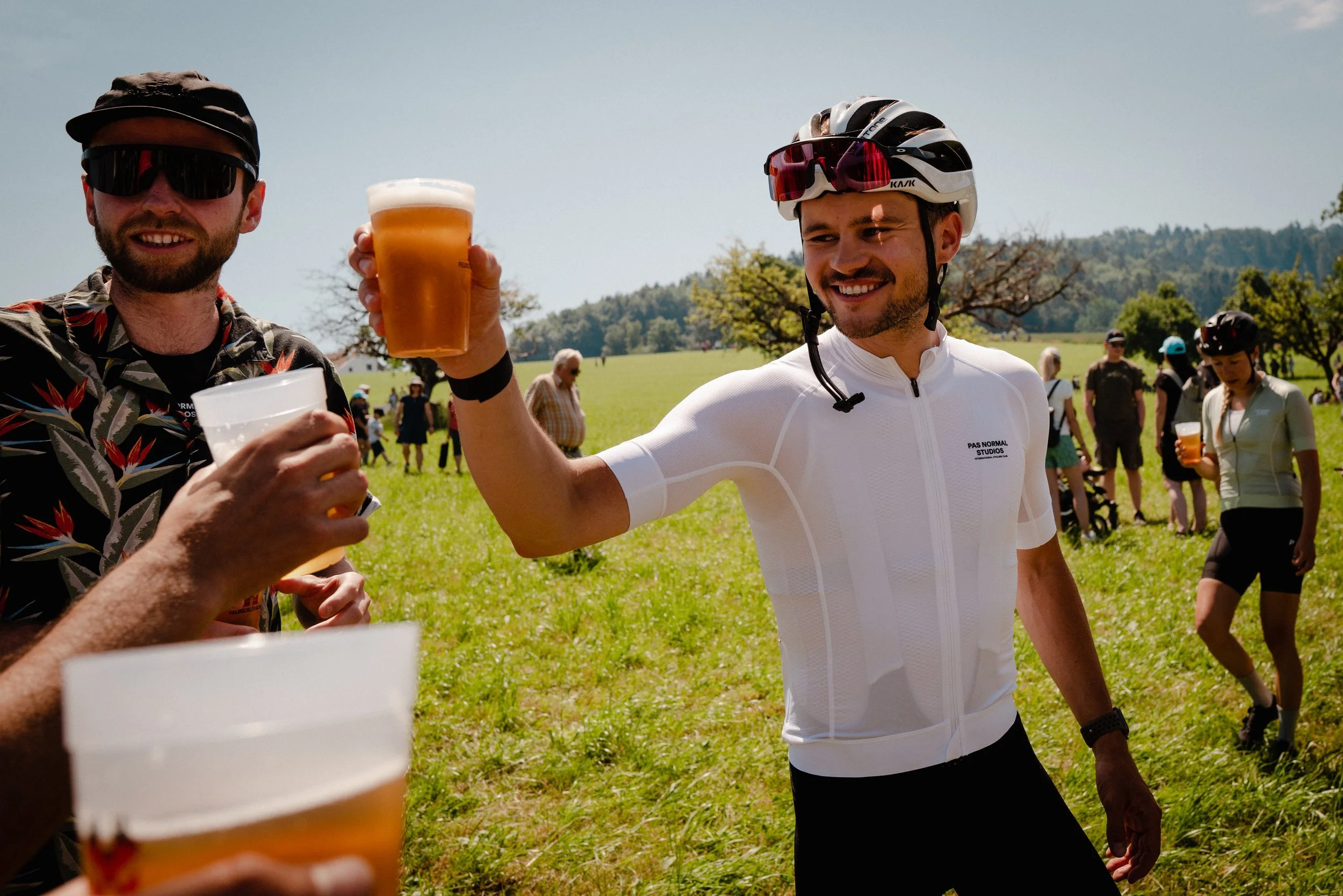Two men raising glasses of beer in a toast outdoors, surrounded by people and trees, some wearing cycling gear and helmets.