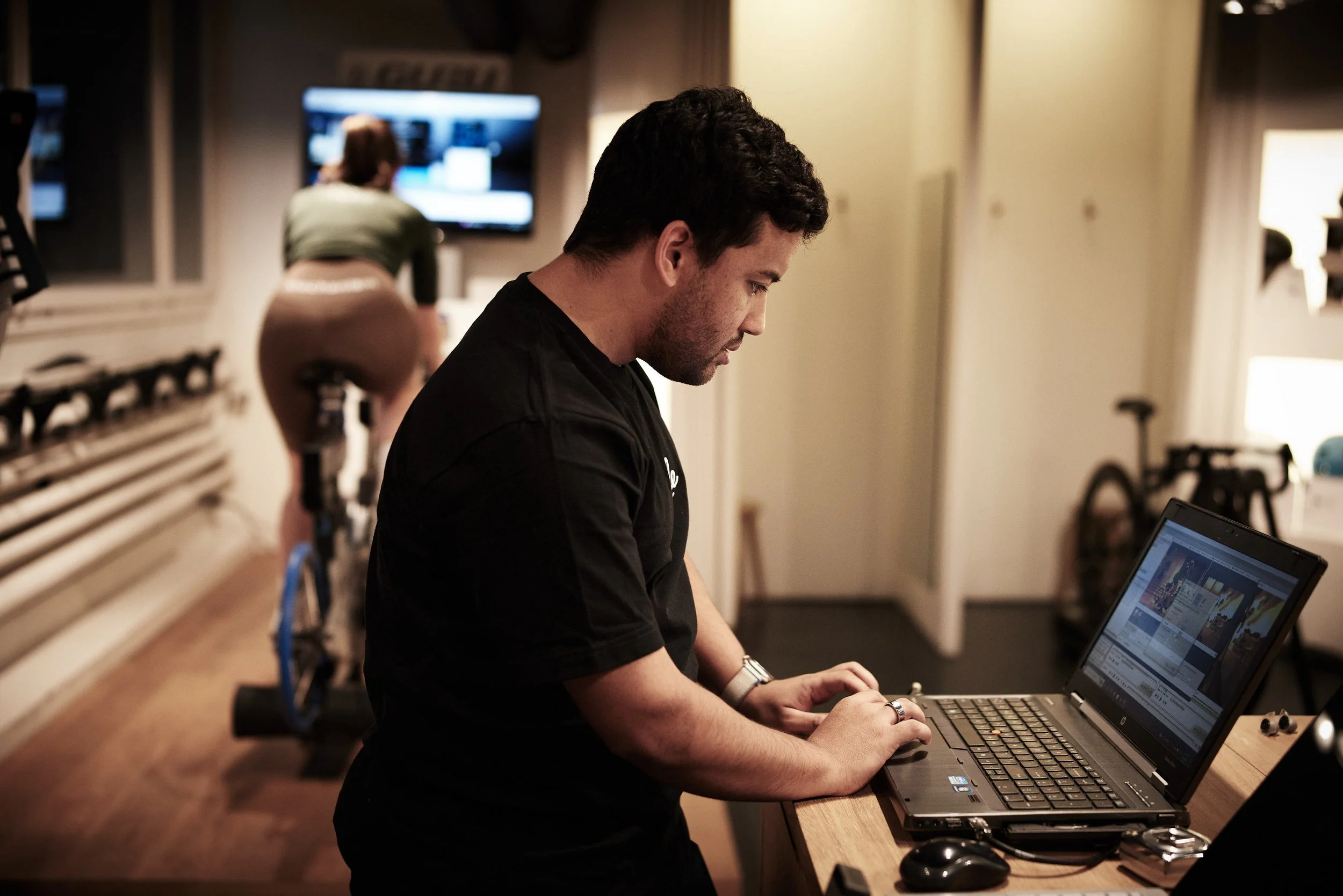 A man working on a laptop at a desk in a gym, with a woman riding a stationary bike in the background.