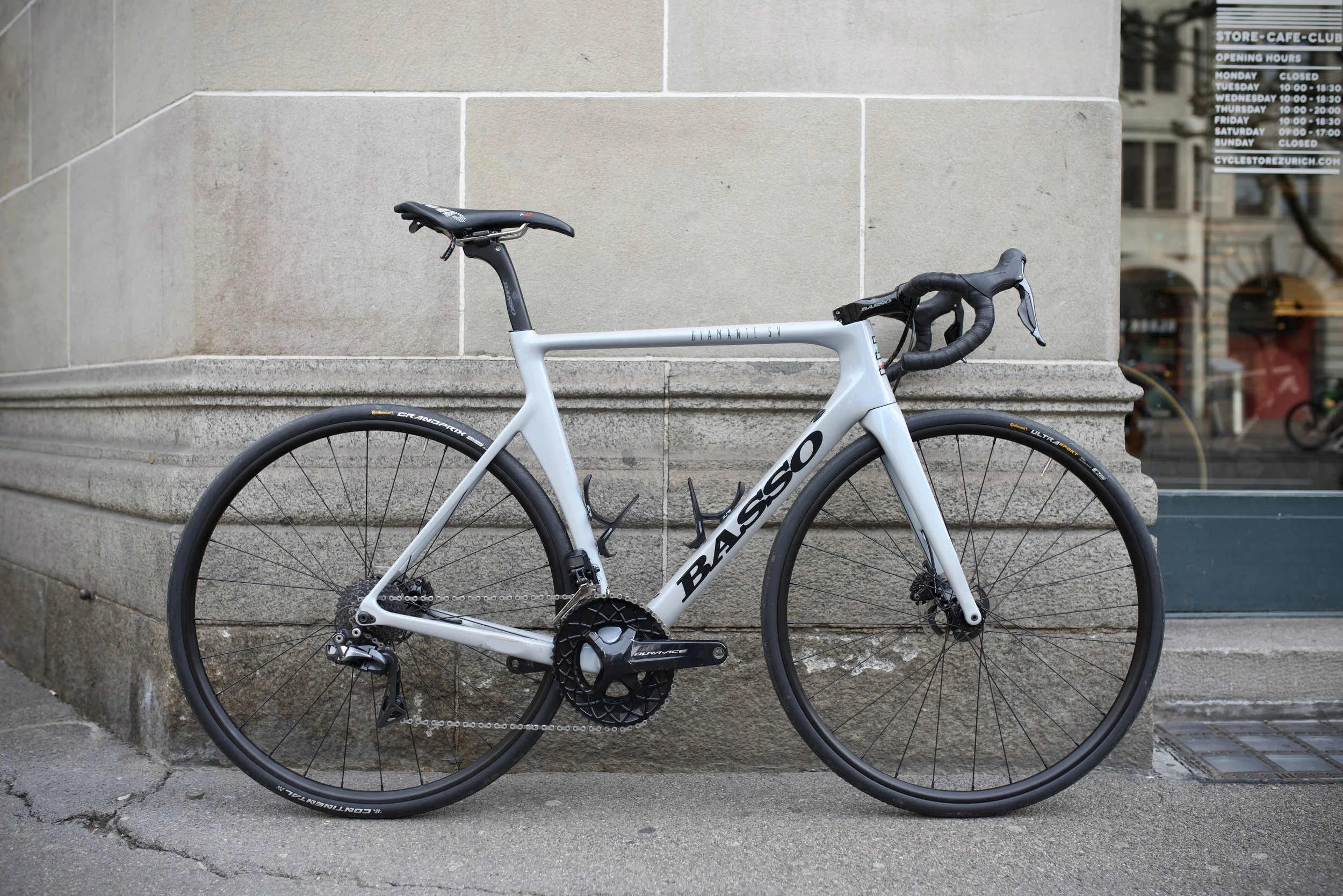A white Basso road bicycle with black tires leaning against a stone wall outside a building with a window and a sign displaying store hours.