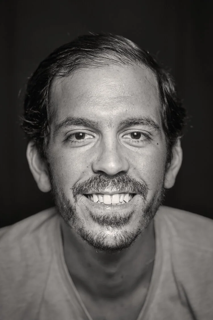 Black and white close-up portrait of a smiling man with short, dark hair, a beard, and mustache, wearing a light-colored shirt against a dark background.