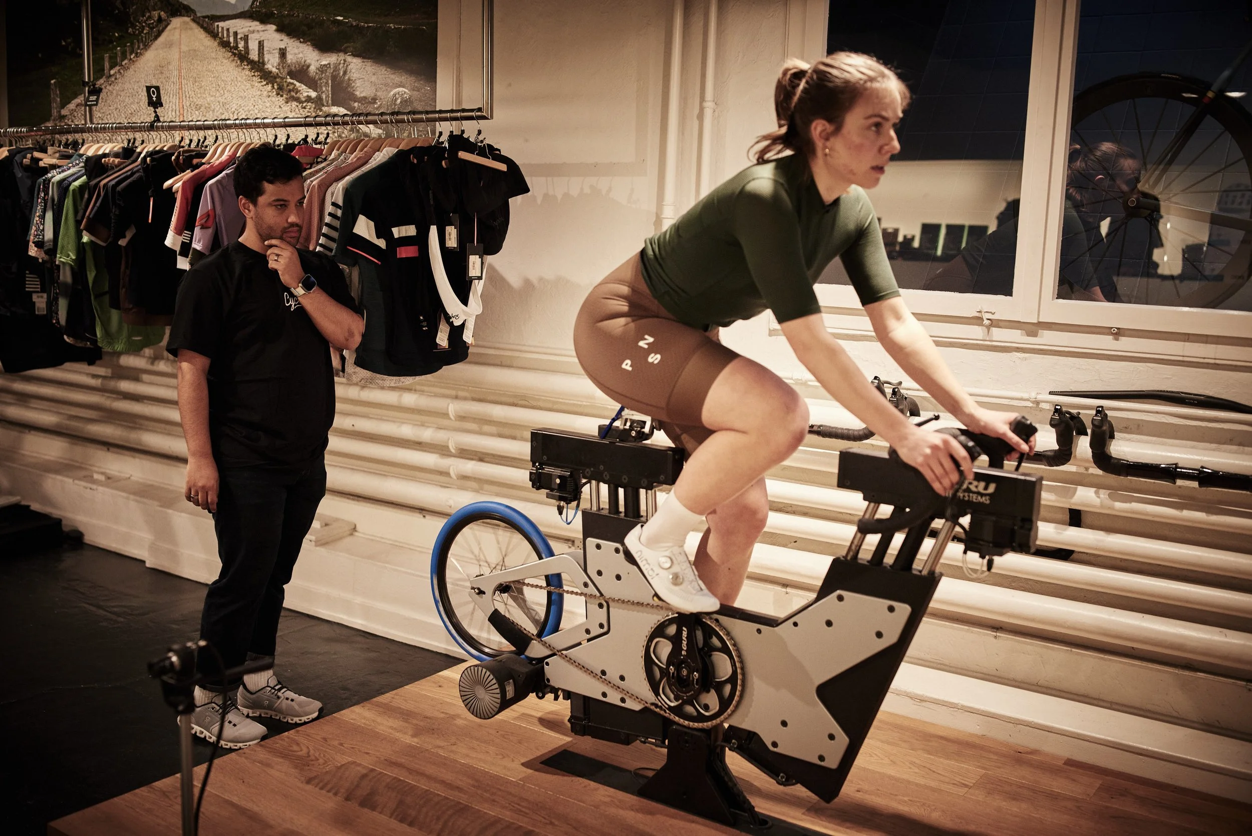 Woman on a stationary exercise bike being monitored by a man in a retail store with sports clothing hanging on the wall.