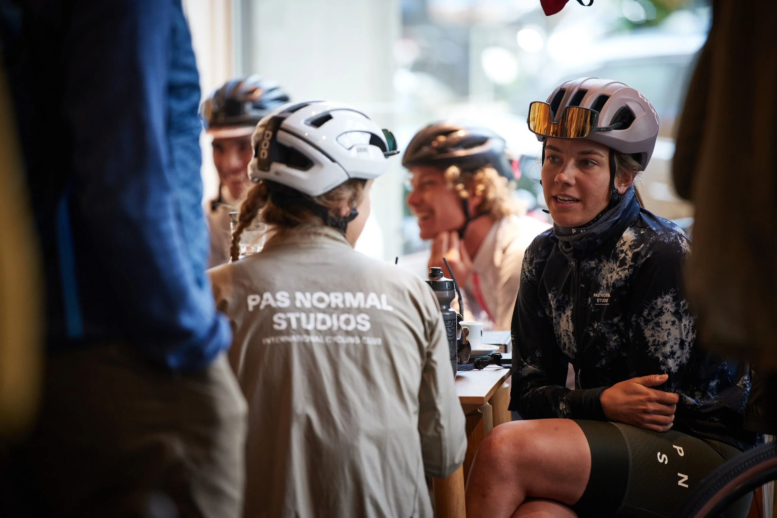 A group of female cyclists wearing helmets and cycling gear sitting and talking inside a cafe, with one woman in black cycling clothes appearing to listen attentively.