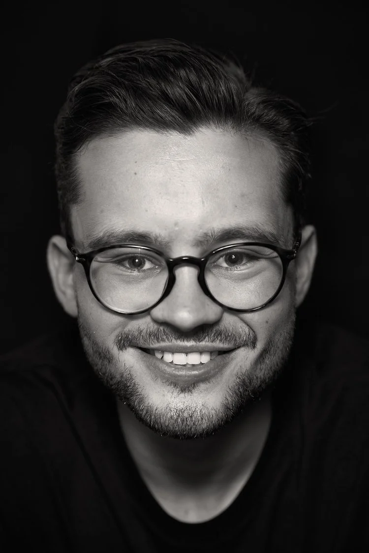 Black and white close-up photo of a young man with glasses, smiling with visible teeth, short styled hair, and a beard.