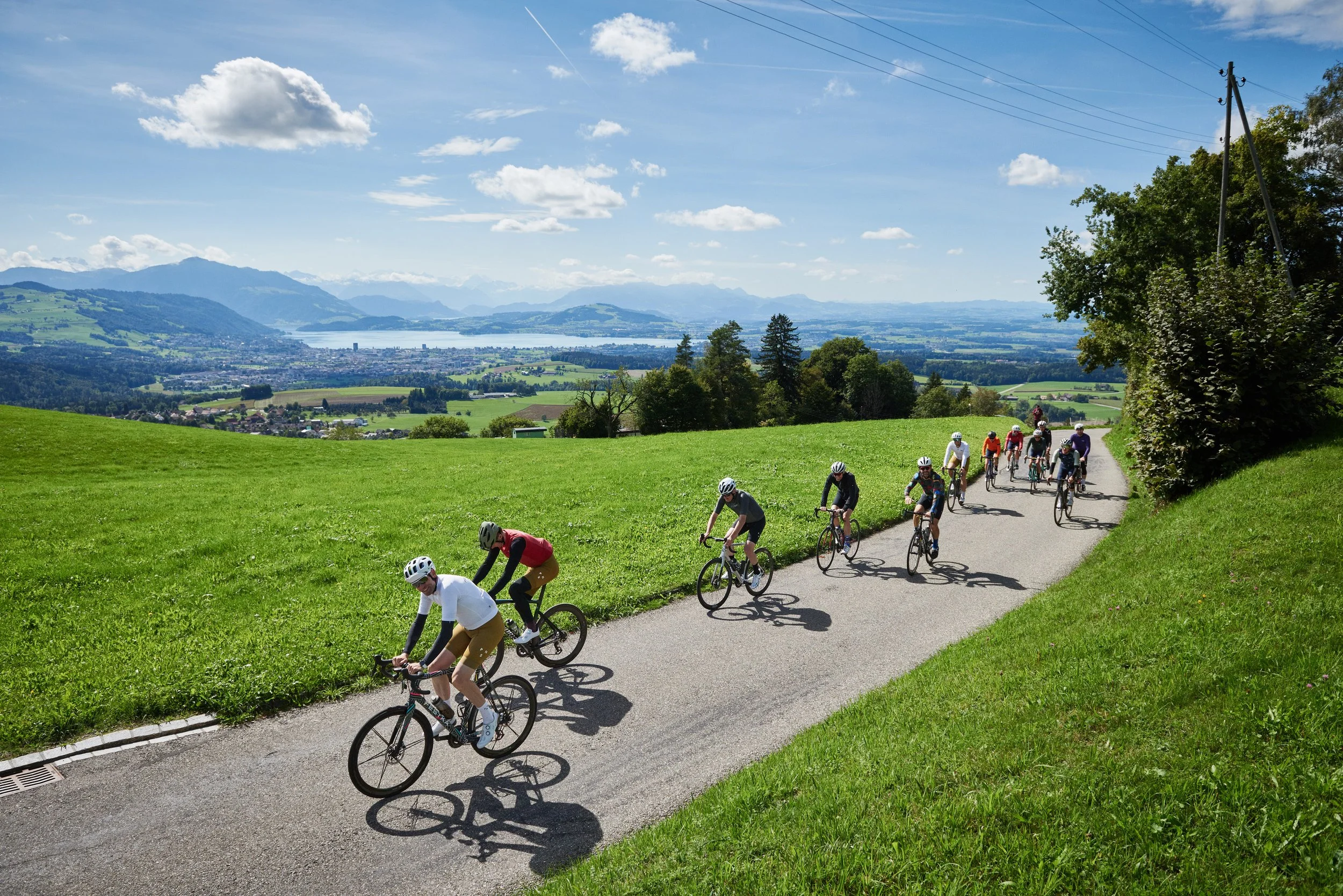 A group of people cycling on a curved road through green fields with mountains and a lake in the background under a partly cloudy sky.