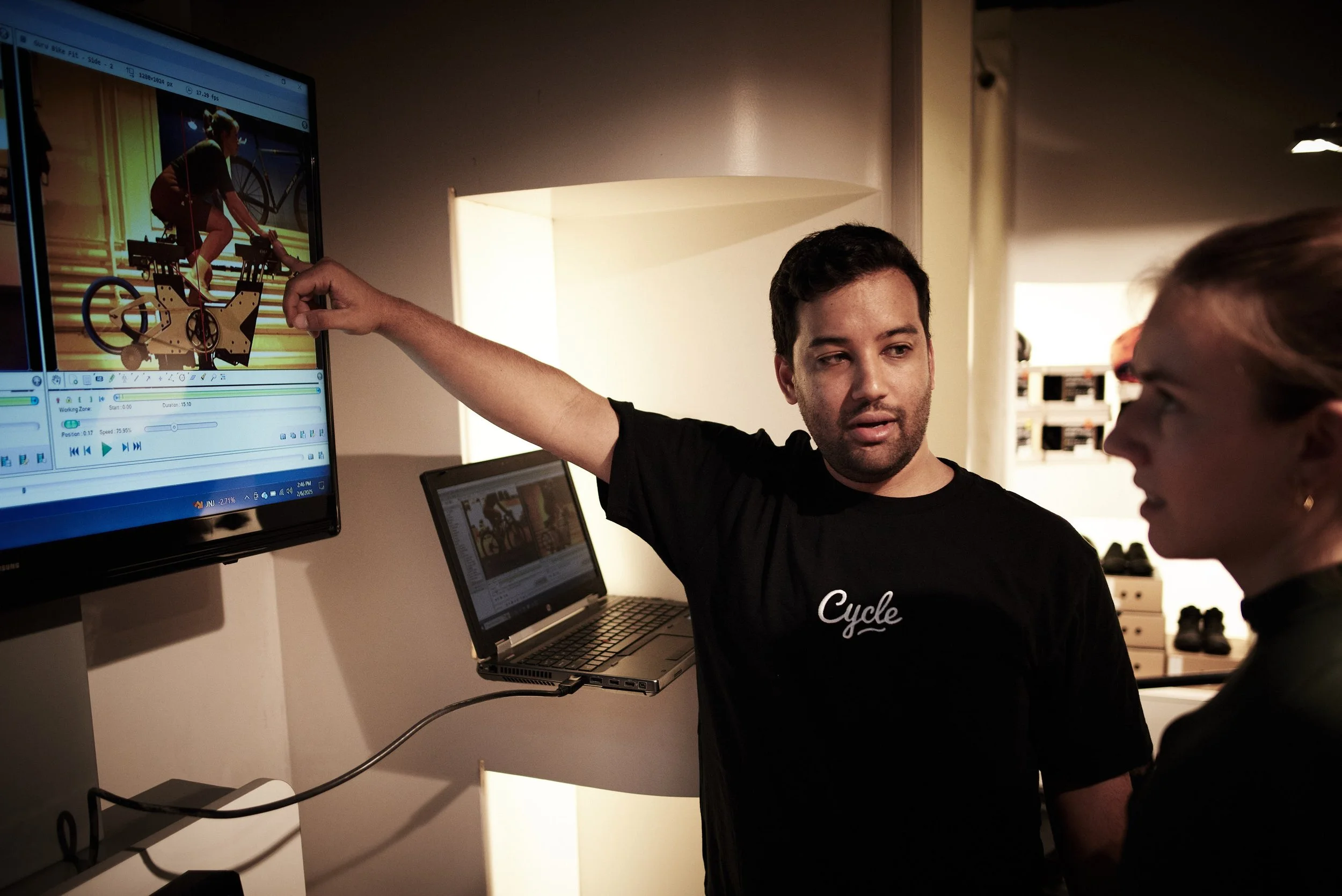 Two people having a discussion, with one person pointing at a computer screen displaying a video of a person on a bicycle simulator.