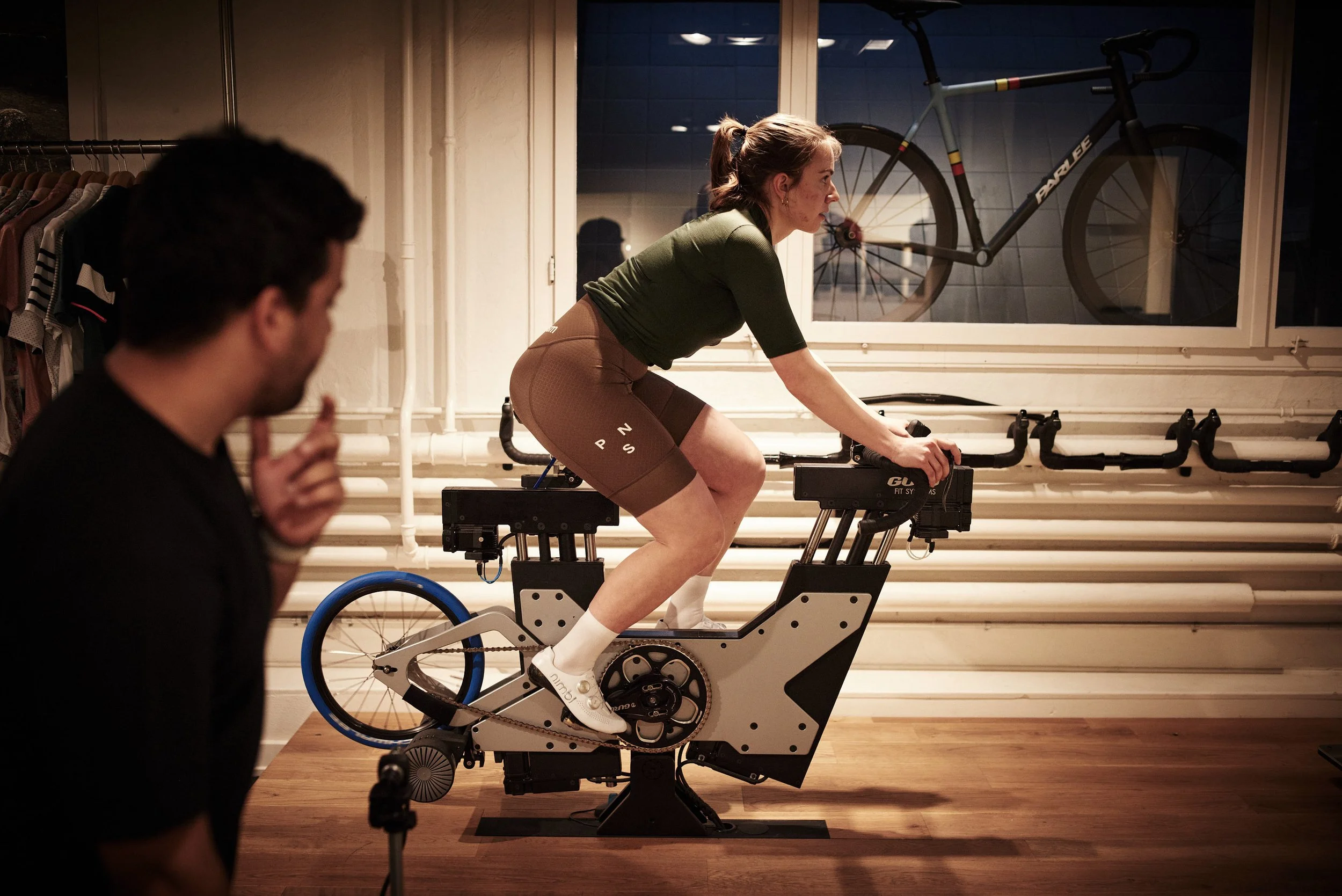 A woman riding an indoor stationary bike with a blue bike visible outside the window, while a man watches and gestures in the foreground.