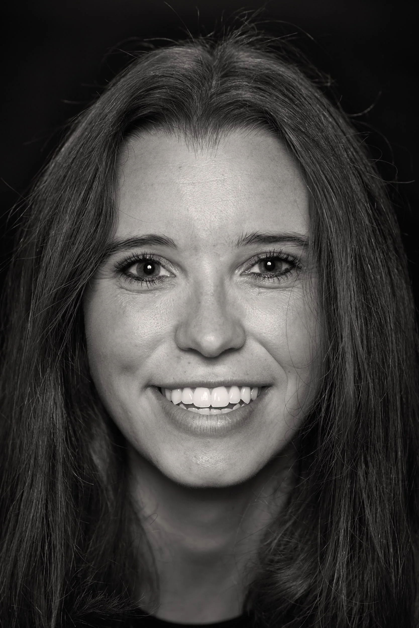 Close-up black and white portrait of a smiling woman with long hair, straight teeth, and bright eyes.