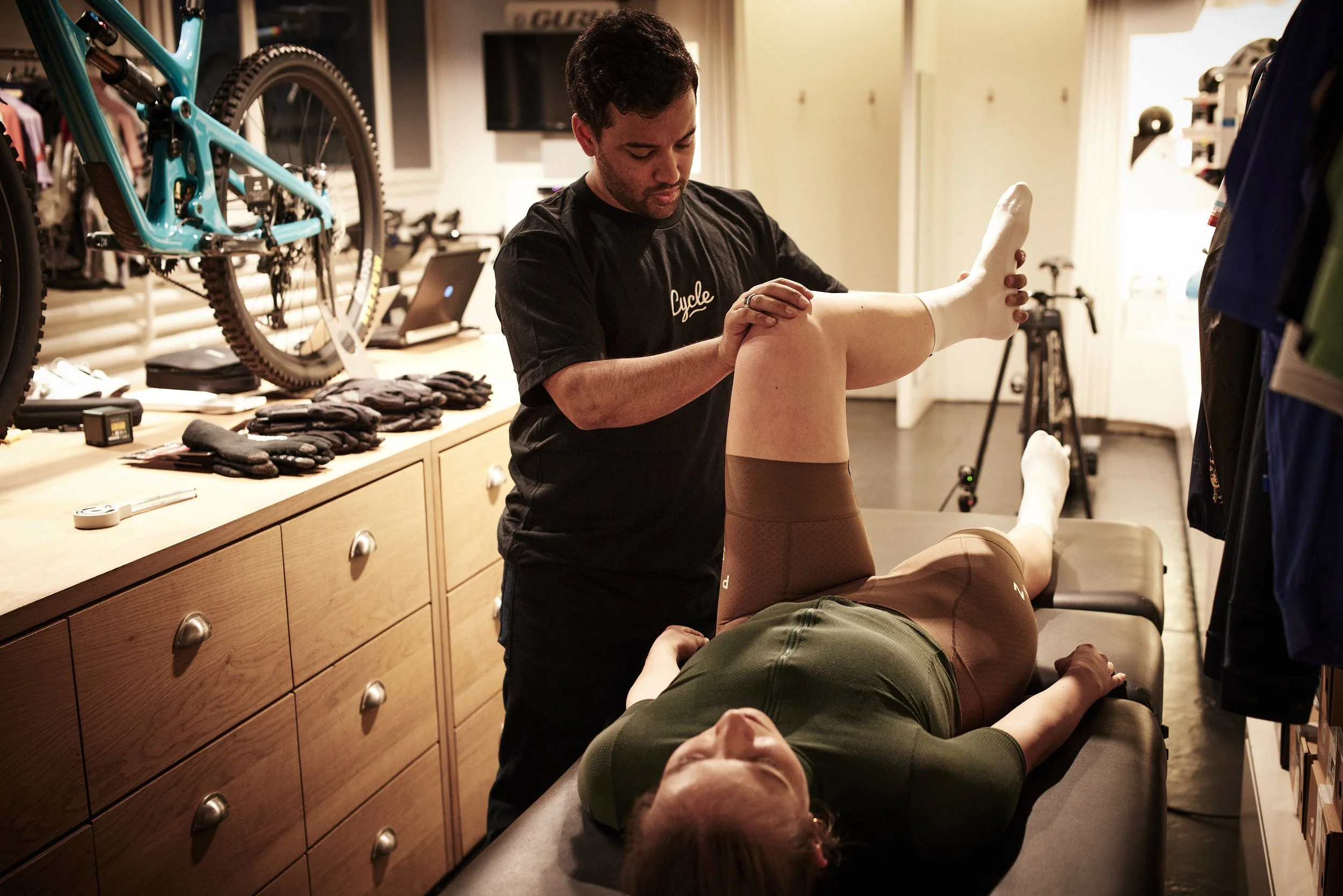 A physiotherapist helps a patient with a leg stretch or exercise on a massage table in a sports or cycling store, with cycling gear and equipment in the background.