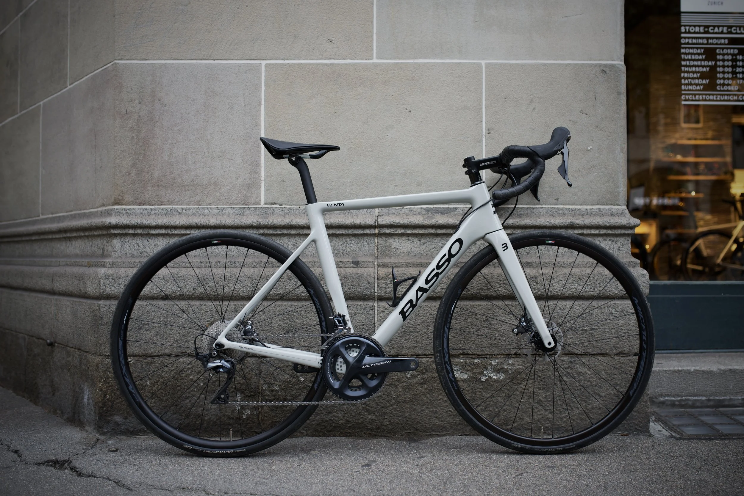 A white Basso road bicycle with black wheels and handlebars parked against a stone building wall exterior.