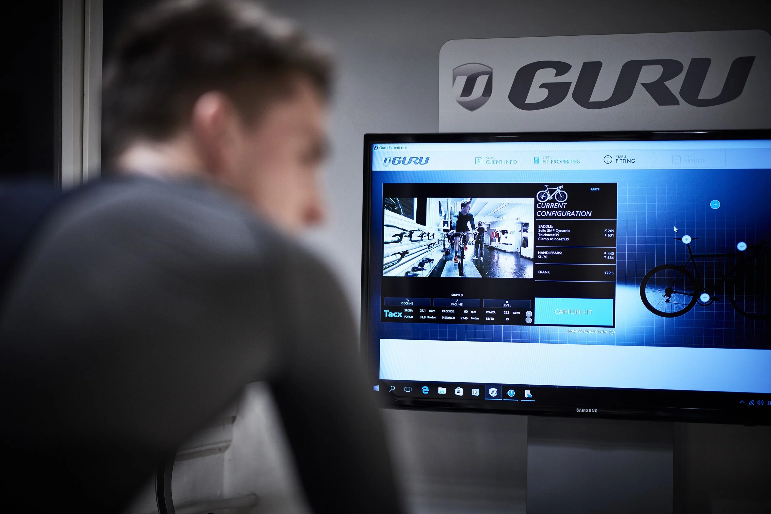 A person in a gray shirt appears to be in a bike fitting studio looking at a computer screen displaying a bike fitting software. The screen shows an image of a person on a bicycle and various technical data.