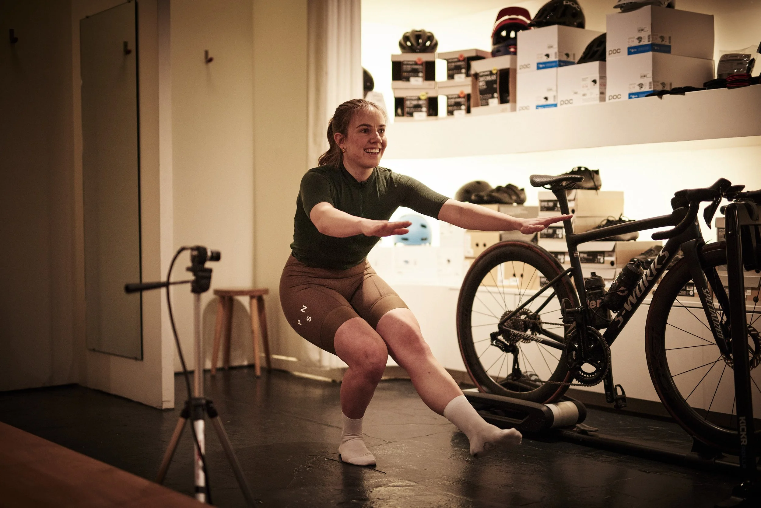 A woman in athletic clothing performs squats in a room with indoor exercise equipment, including a stationary bike and a camera on a tripod.