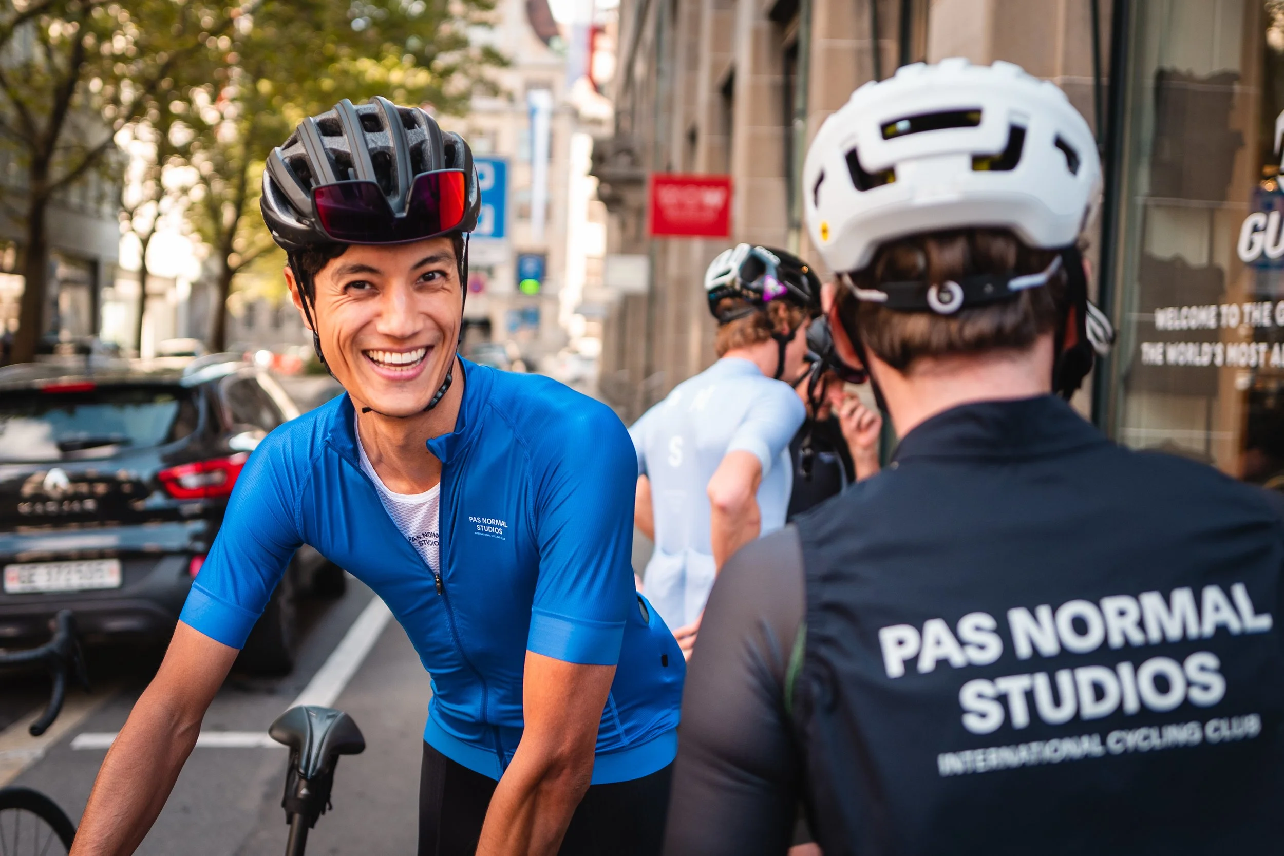 Smiling cyclist in blue jersey talking to another cyclist wearing a black jersey with white text, on a city street with cars and buildings in the background.