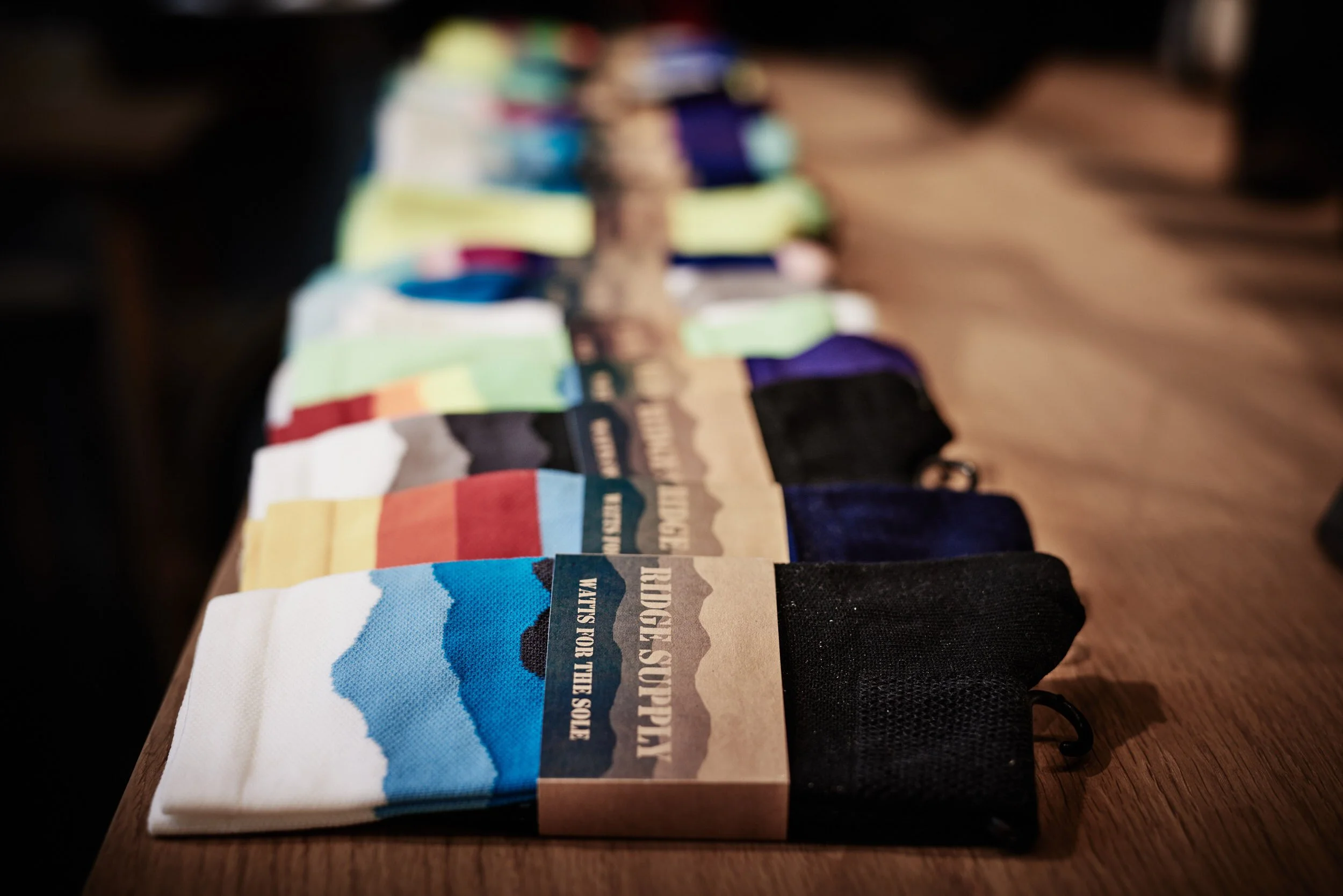 A row of colorful socks with mountain designs and black tops, arranged on a wooden table, each sock wrapped with a paper band reading "WILDERNESS SOCKS - SOLES FOR THE SOUL".