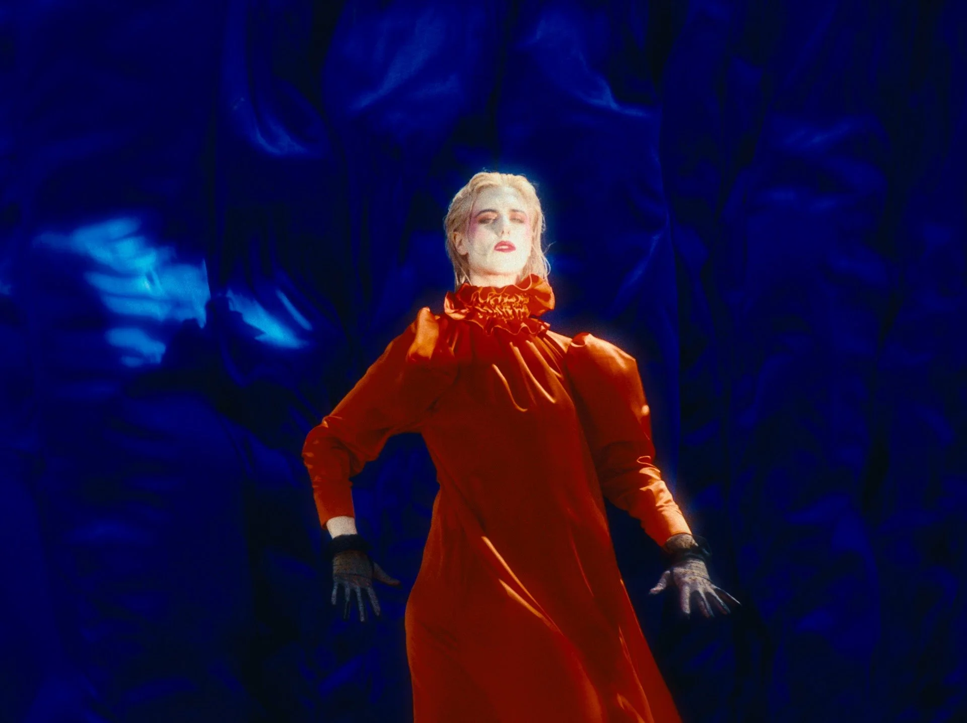 A woman with red lipstick and a ruffled orange dress poses with her left hand, wearing a black fishnet glove, covering her eyes from sunlight.
