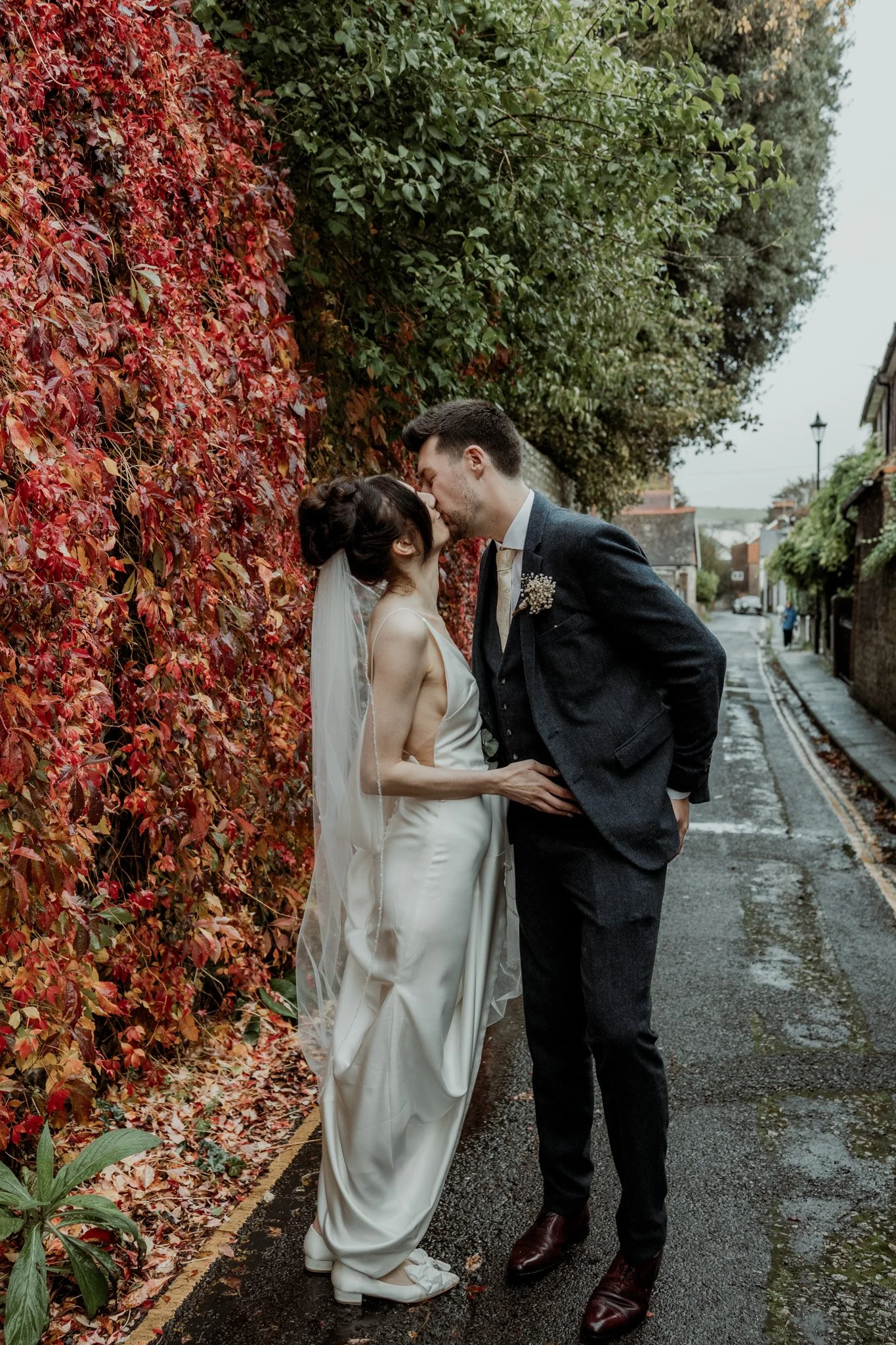 A bride and groom kissing on a wet street. The bride is wearing a white wedding dress and veil, and the groom is in a dark suit. They are standing next to a wall covered in vibrant red and green leaves.