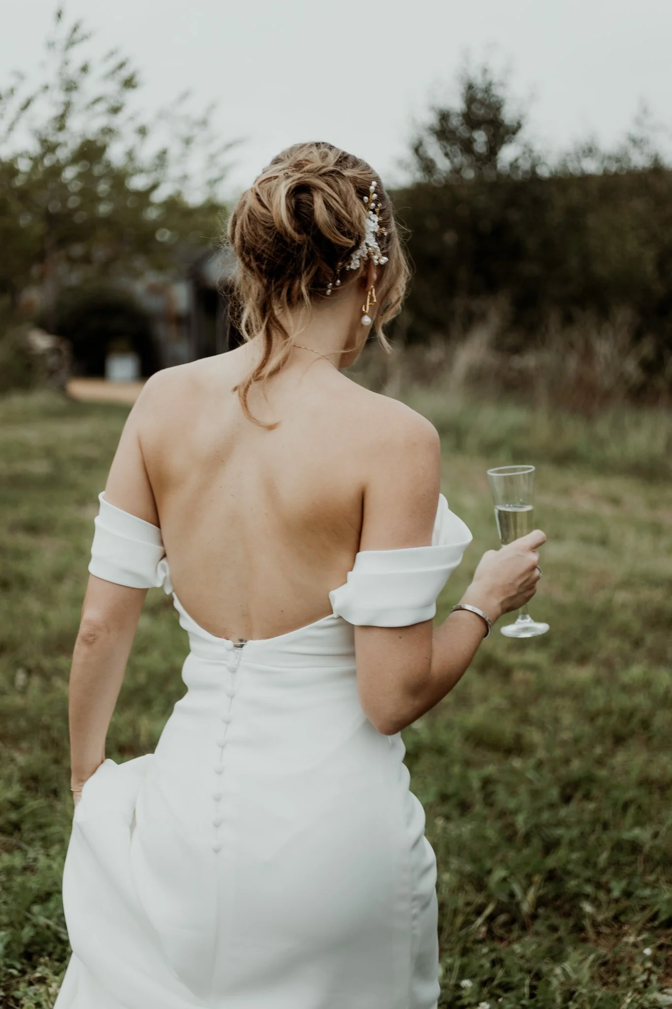 Bride in open-back wedding dress holding champagne glass, standing outdoors.