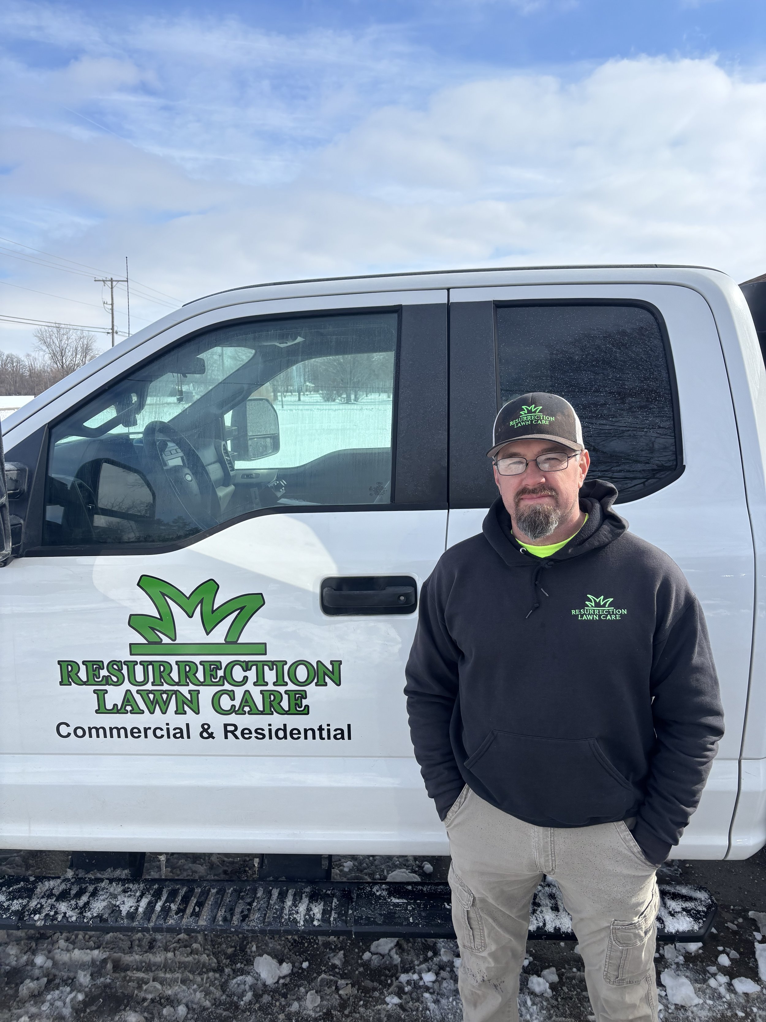 Man standing next to a white Resurection Lawn Care truck wearing a black hoodie and a baseball cap, on a snowy day.