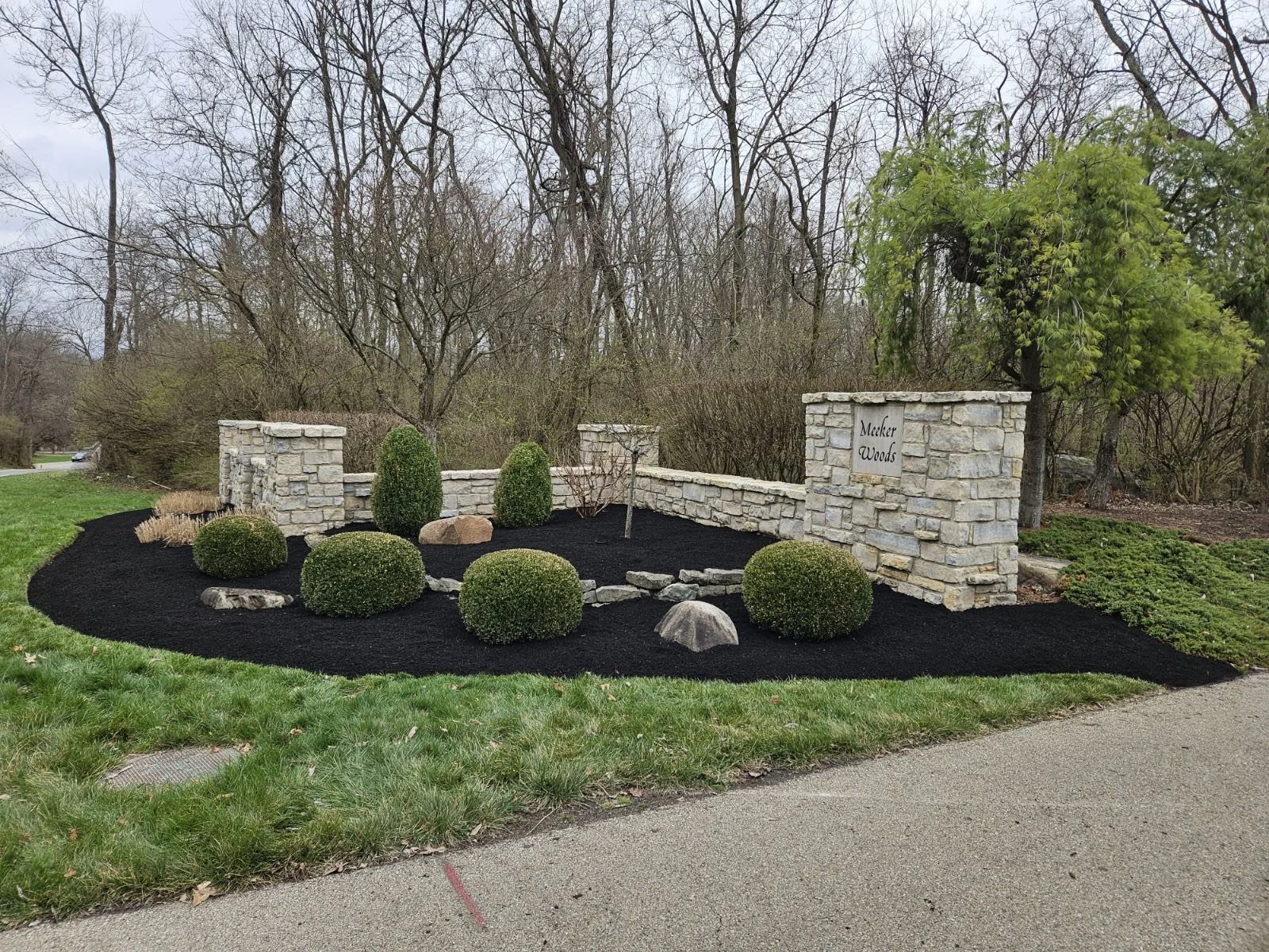 A landscaped garden area with a stone wall and greenery, featuring manicured bushes, mulch, and trees, with a sign reading "Meker Woods."