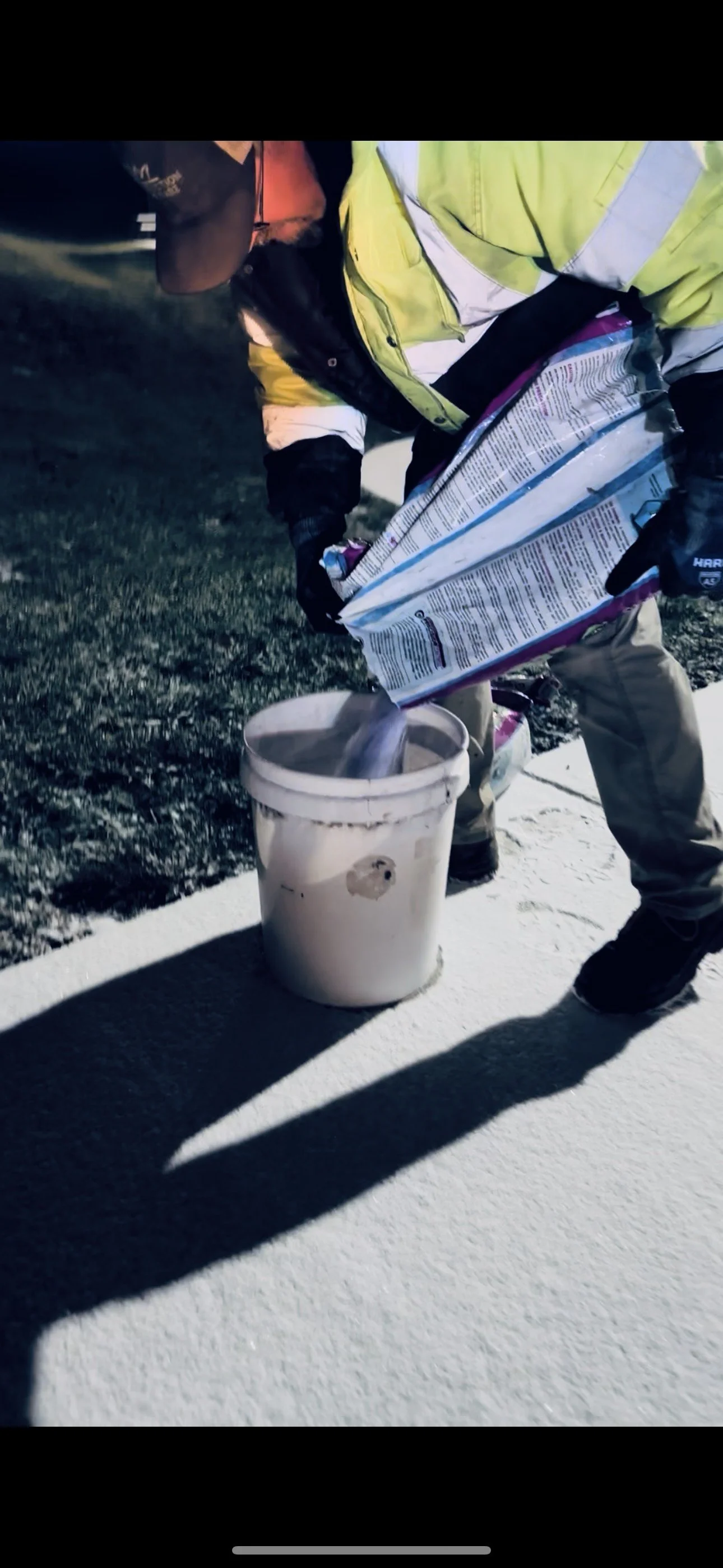Worker in high-visibility jacket pours salt or de-icing material from a bag into a bucket on an outdoor pavement.