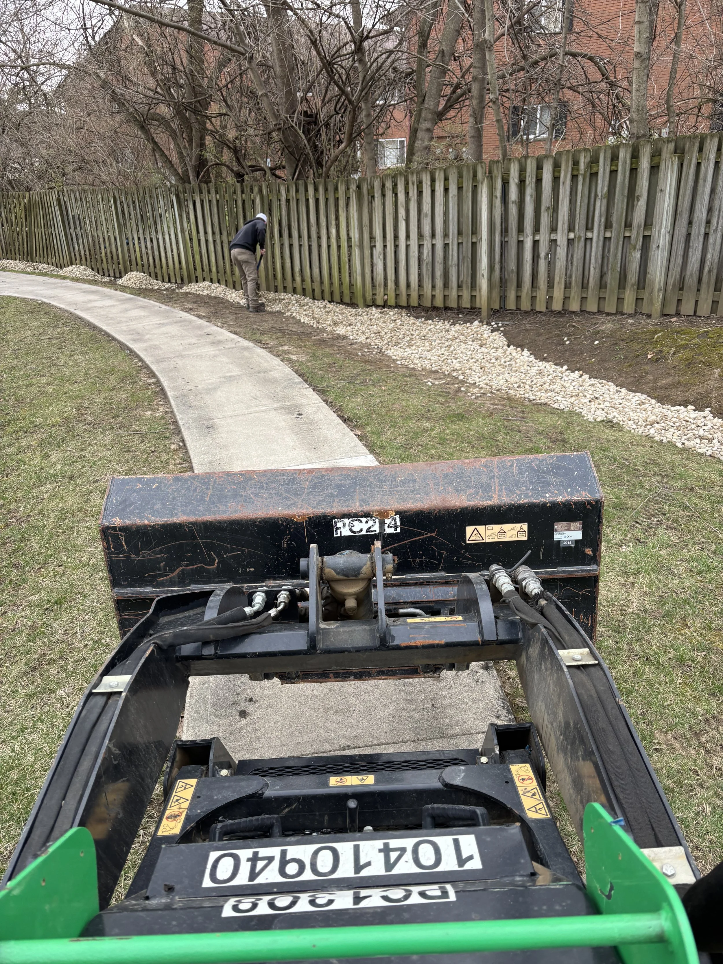 A person is working along a garden bed near a wooden fence, with a small construction vehicle in the foreground.