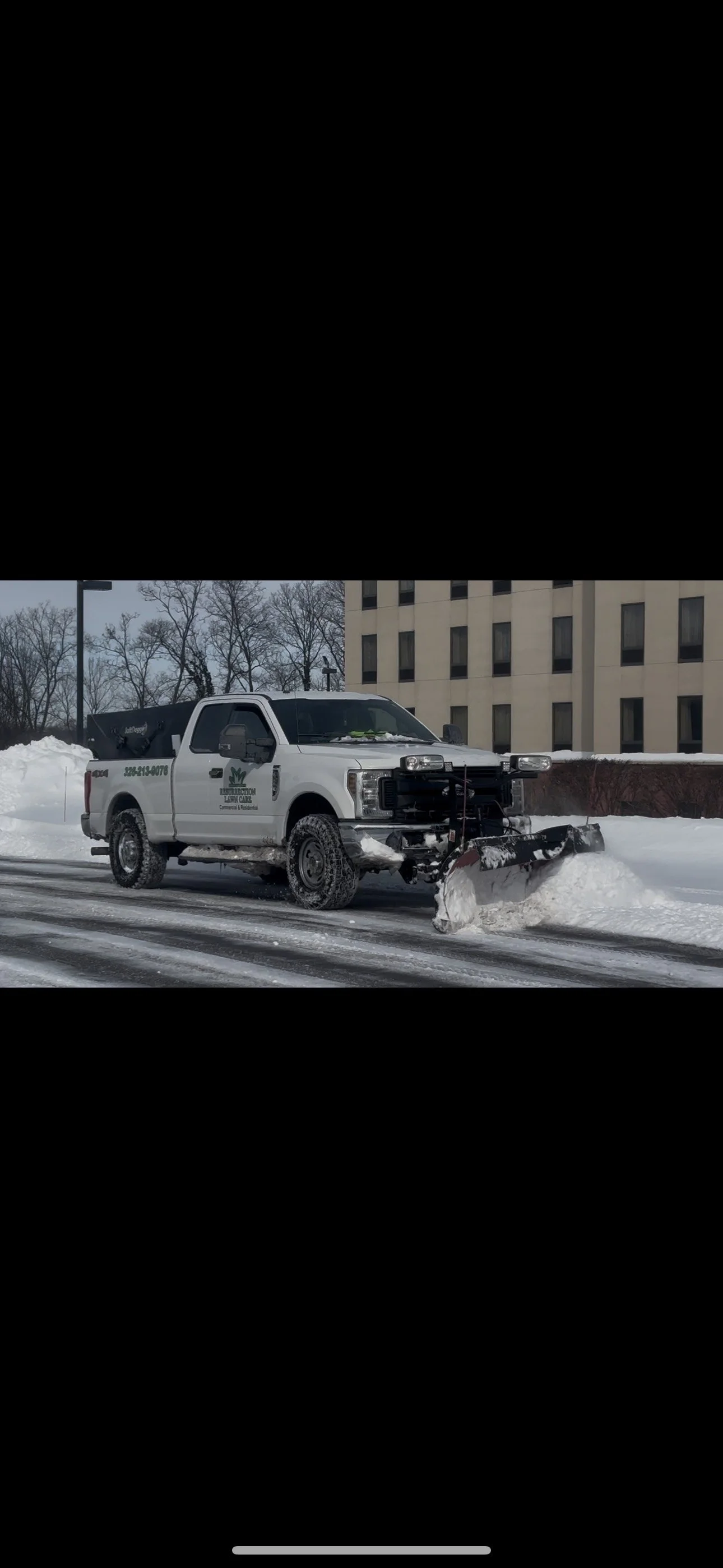 A white snow plow truck clearing snow from a road in winter, with a building and leafless trees in the background.