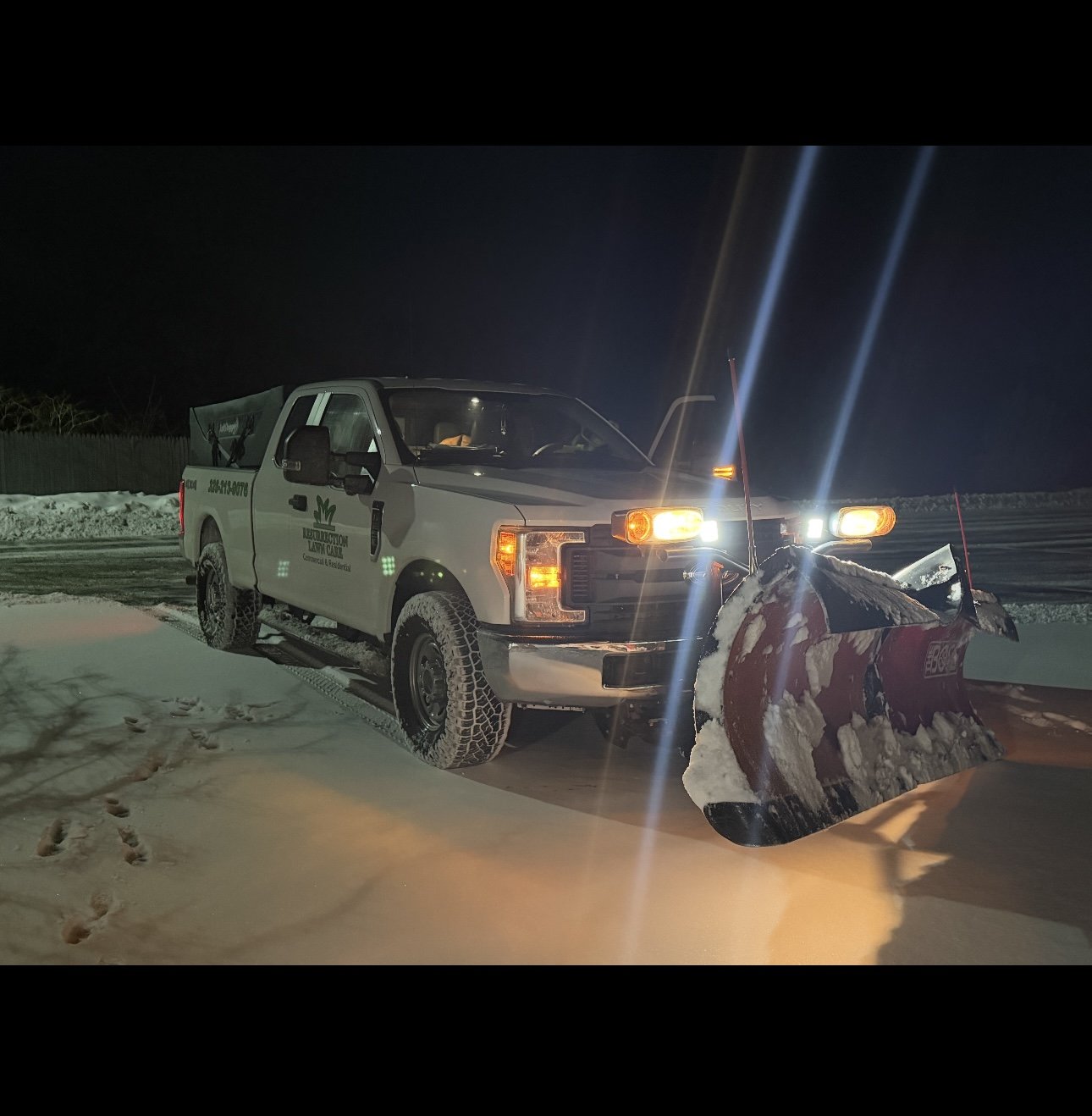 A black pickup truck equipped with a snowplow blade attached to the front, clearing snow on a snowy road surrounded by snow-covered trees.