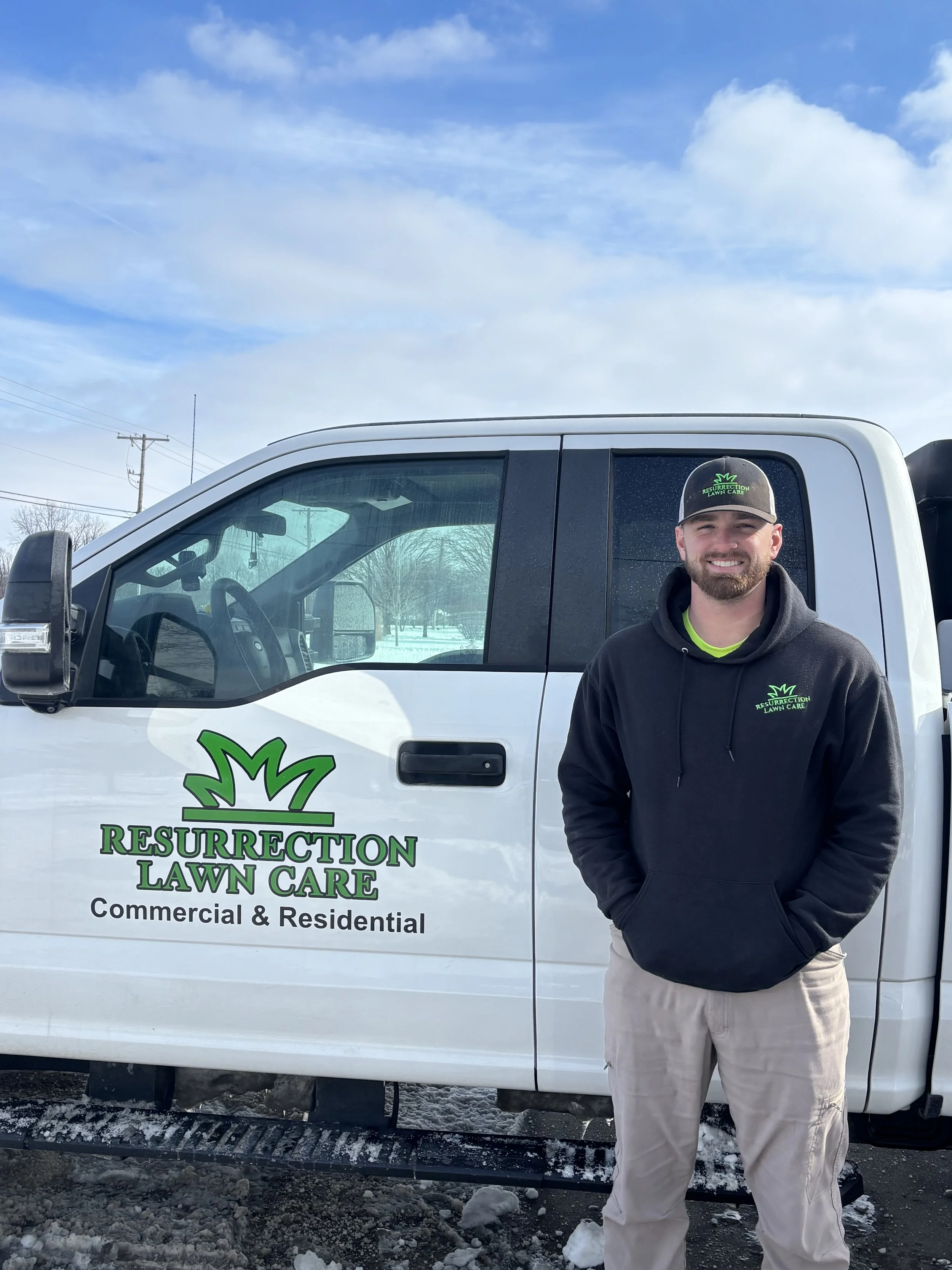 Man standing in front of a white Resurection Lawn Care truck, wearing a black hoodie and a matching cap, smiling outdoors with a snowy ground and cloudy sky.